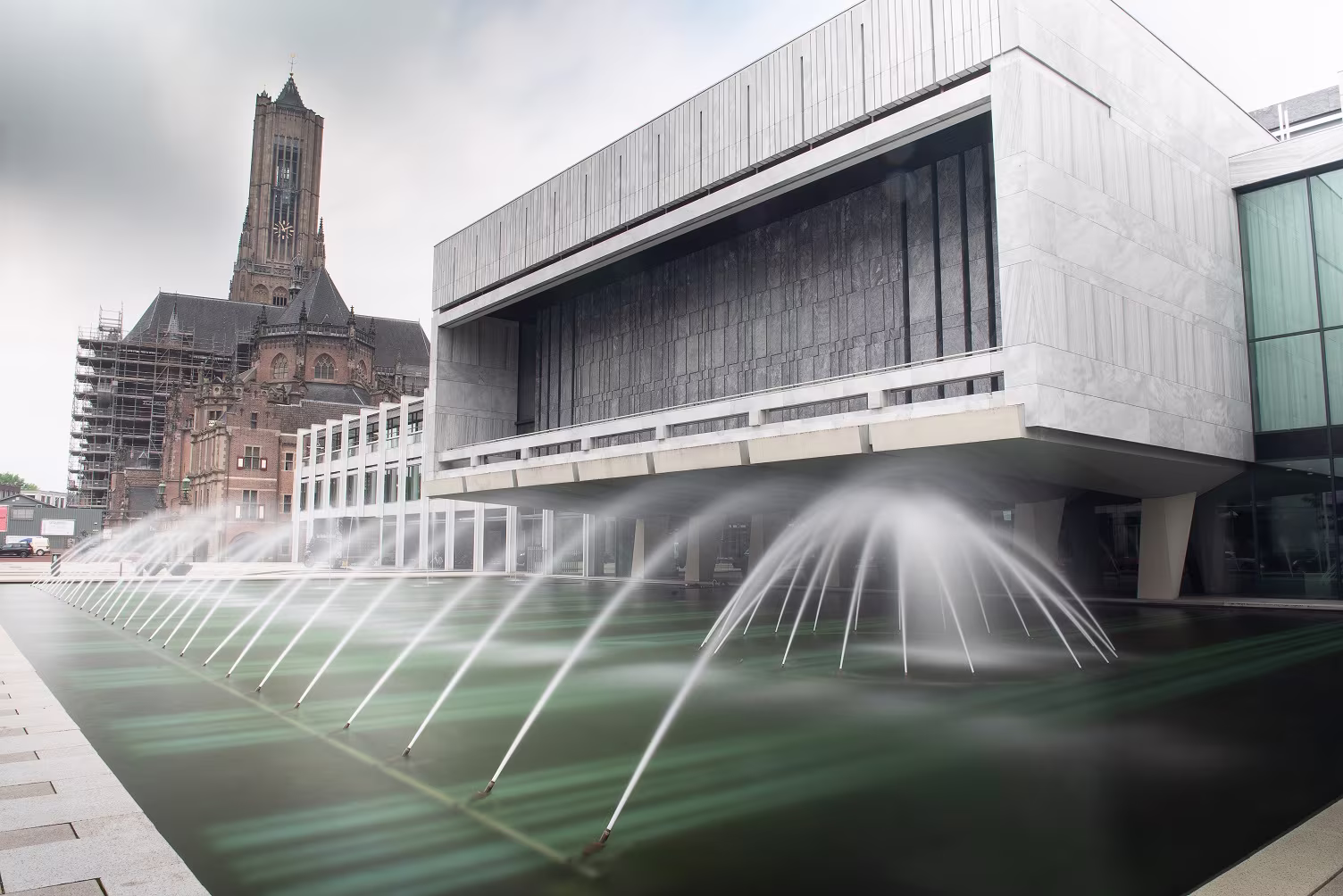 Modern gemeentehuis van Arnhem met fontein op voorgrond en historische kerktoren op achtergrond onder bewolkte hemel.
