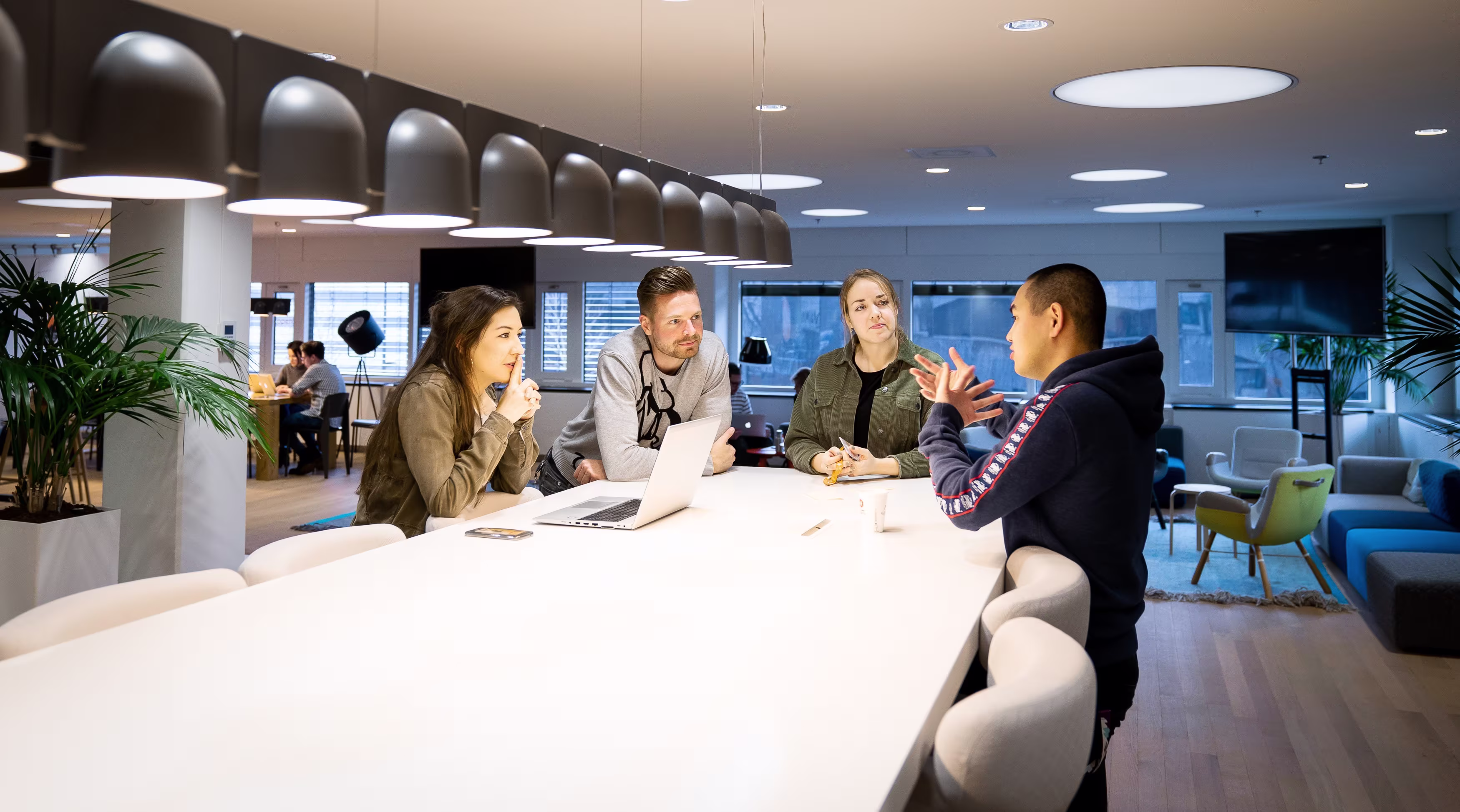 Vier collega's zitten rond een witte tafel in een modern kantoor en voeren een gesprek tijdens een werkoverleg.
