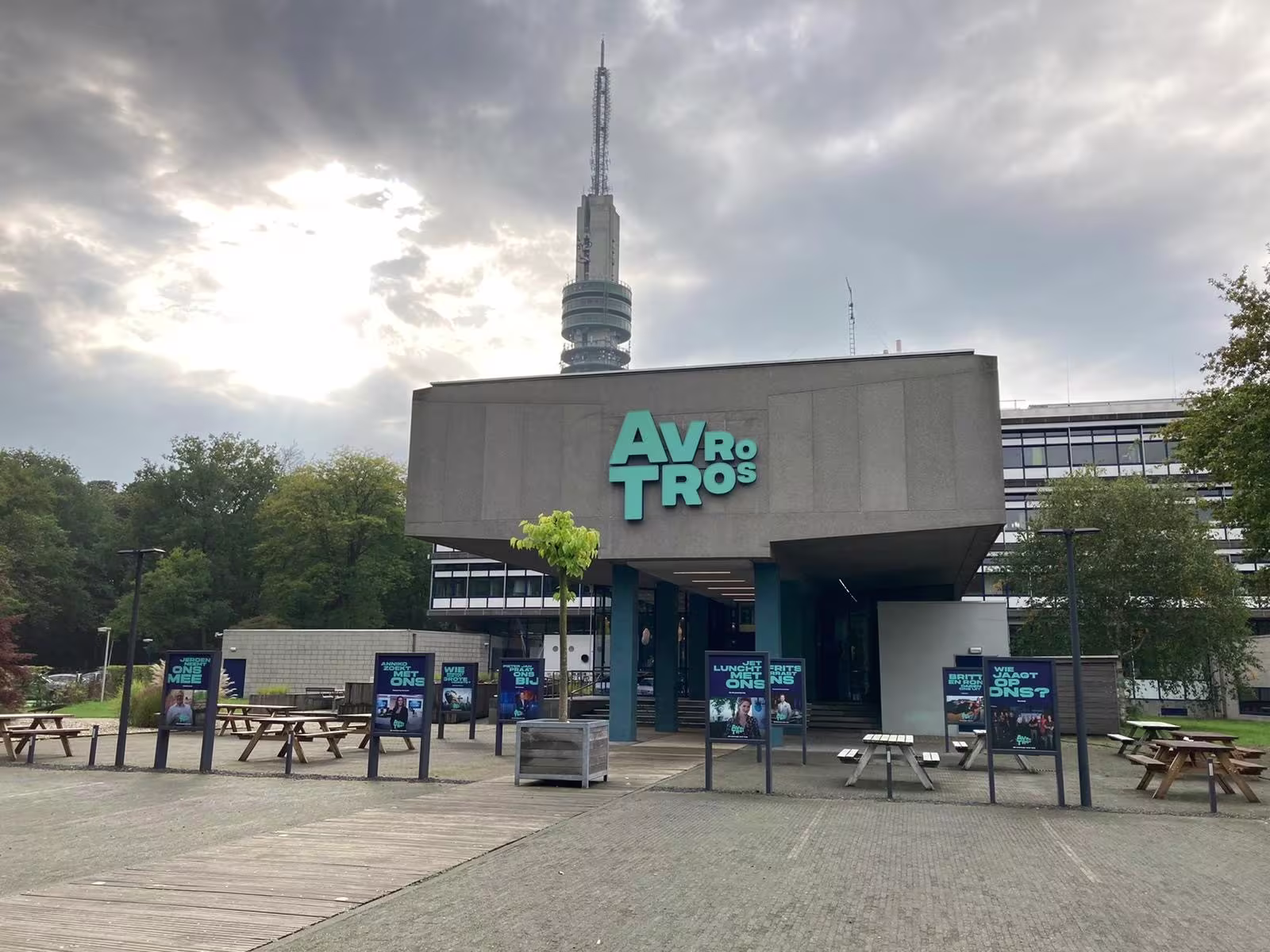 Het AVROTROS-gebouw met logo op de gevel, omgeven door groen en met een zendmast op de achtergrond onder een bewolkte lucht.