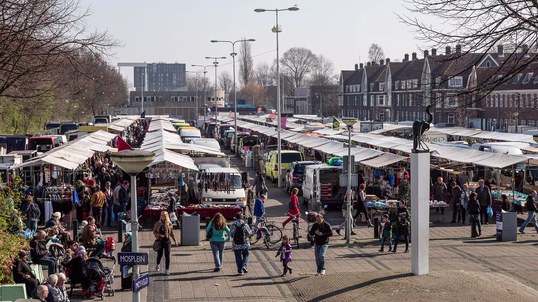 Een drukke markt met kraampjes en bezoekers op een plein, omringd door typisch Nederlandse huizen en moderne gebouwen.