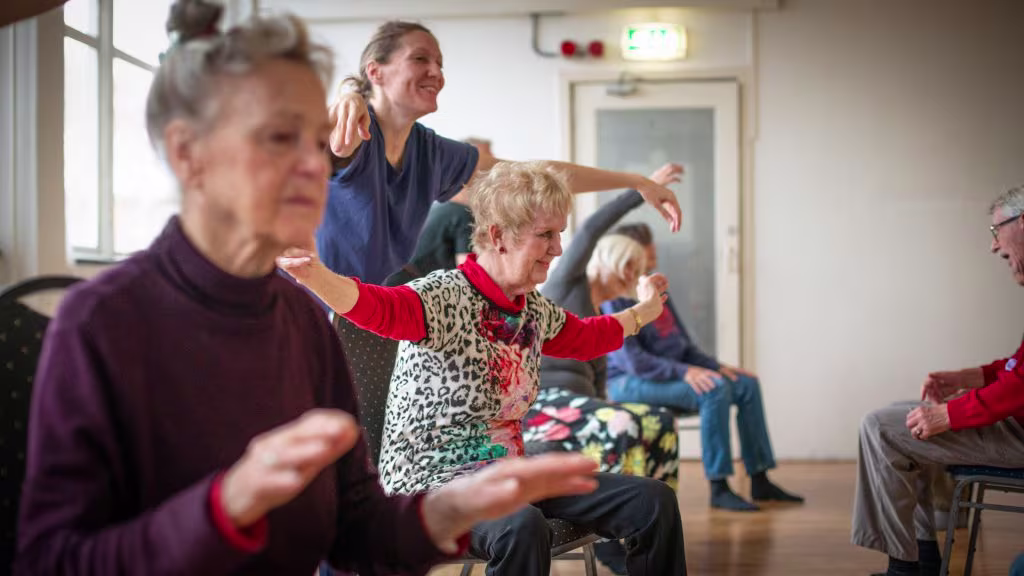 Ouderen doen bewegingsoefeningen onder begeleiding van een zorgverlener in een lichte ruimte.