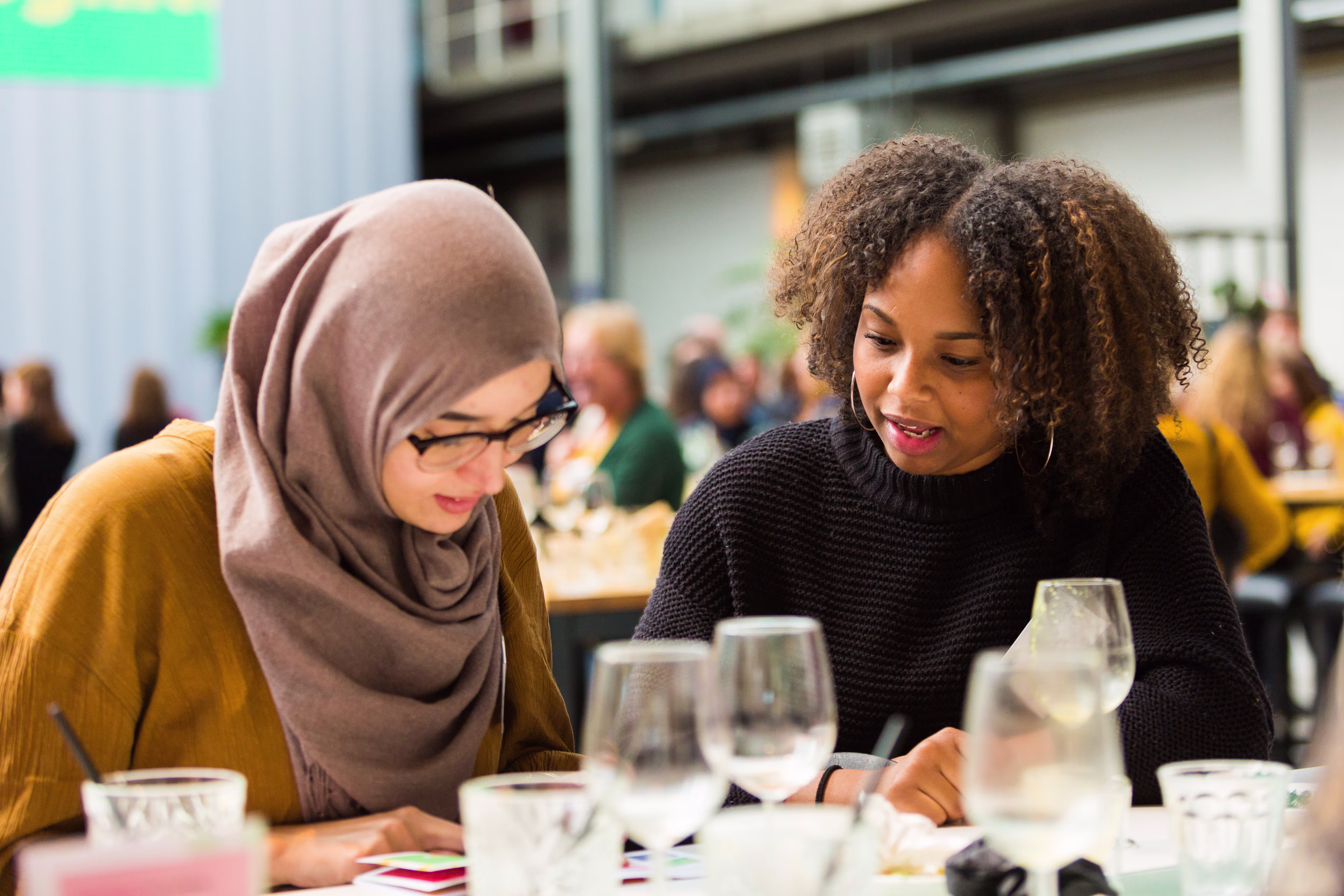 Twee vrouwen zitten aan tafel en bekijken samen documenten, met glazen en andere mensen op de achtergrond in een restaurant.