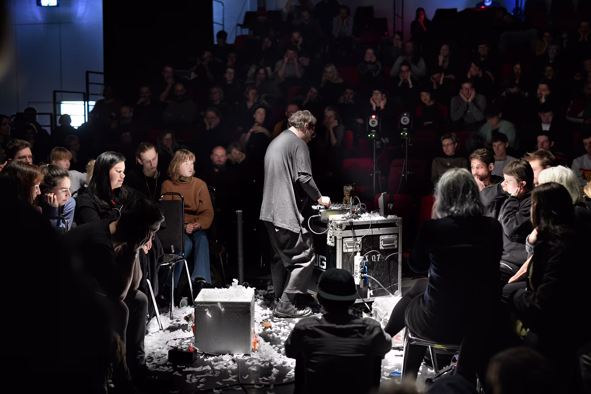 Een performer staat bij geluidsapparatuur omringd door een publiek in een intieme, donkere concertzaal tijdens een experimentele muziekvoorstelling.
