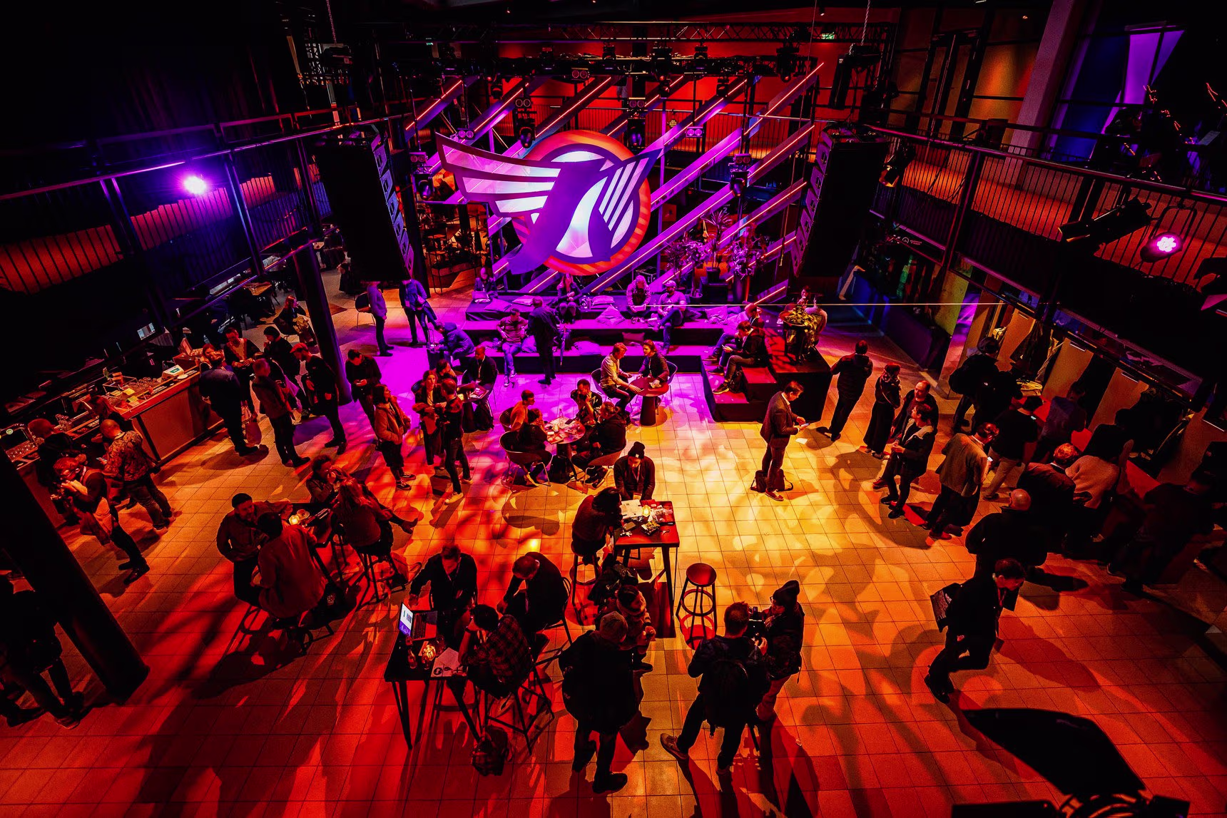Een overzicht van een muziekfestival met paarse en oranje verlichting, waar bezoekers rondlopen in een grote evenementenhal.