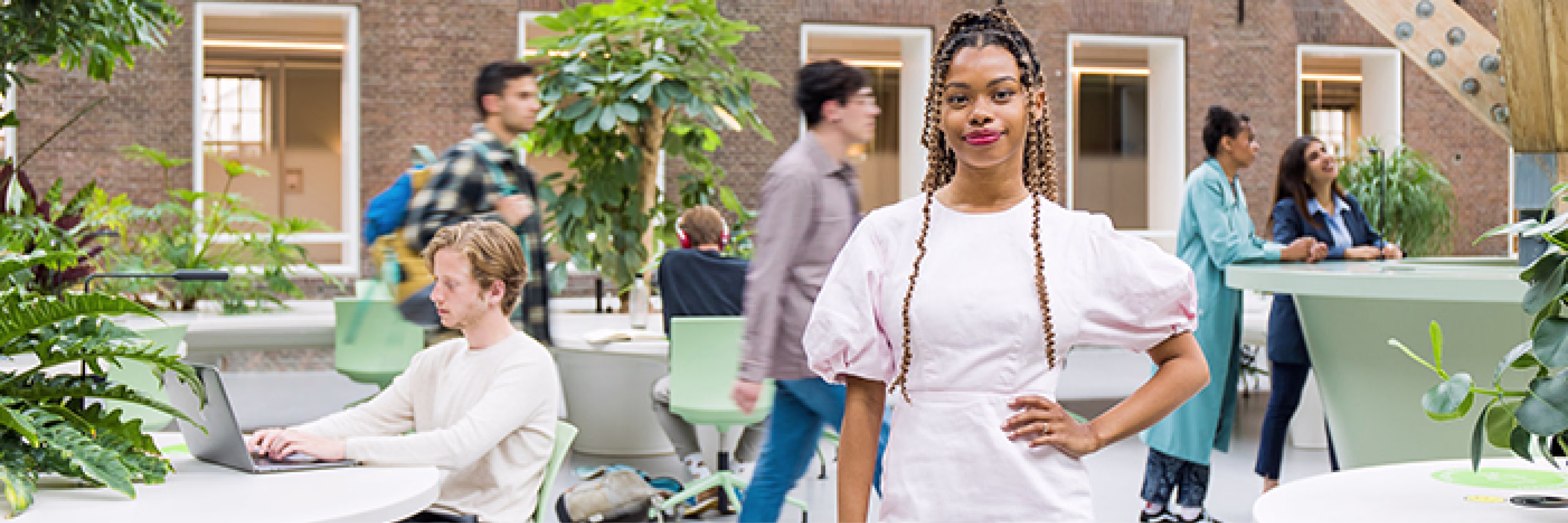 Vrouw in witte blouse staat zelfverzekerd in moderne werkruimte met collega's die werken en overleggen op de achtergrond.