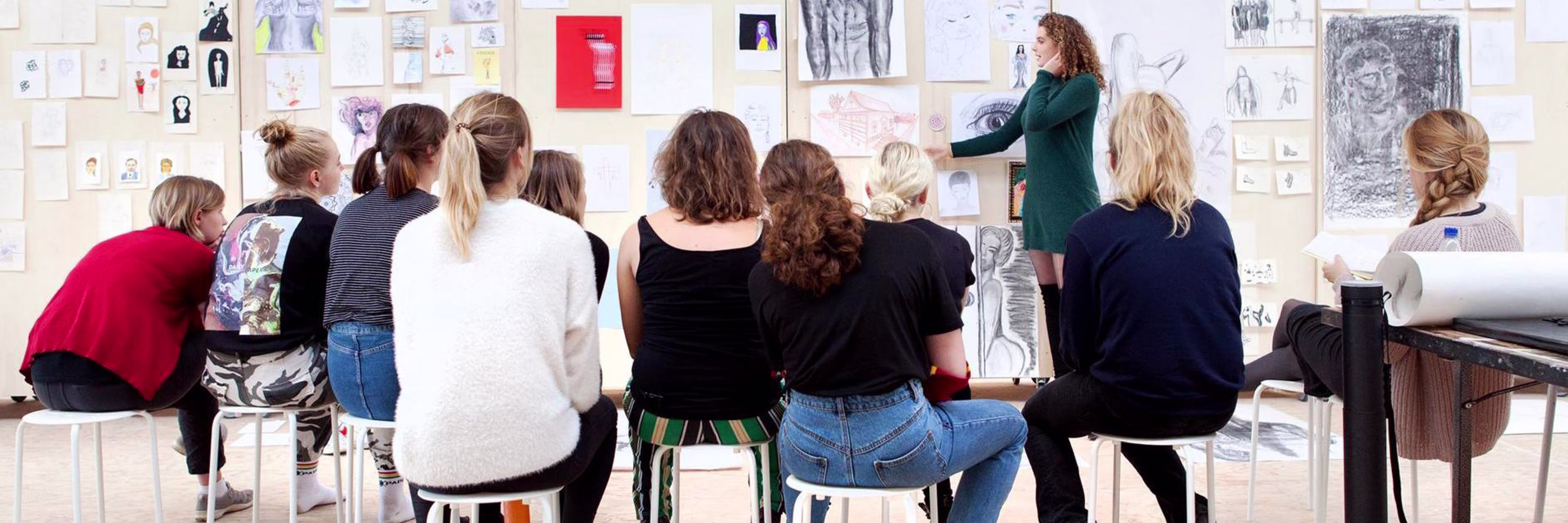 Een docent geeft les aan studenten die op krukken zitten voor een muur vol kunstwerken en tekeningen in een atelier.