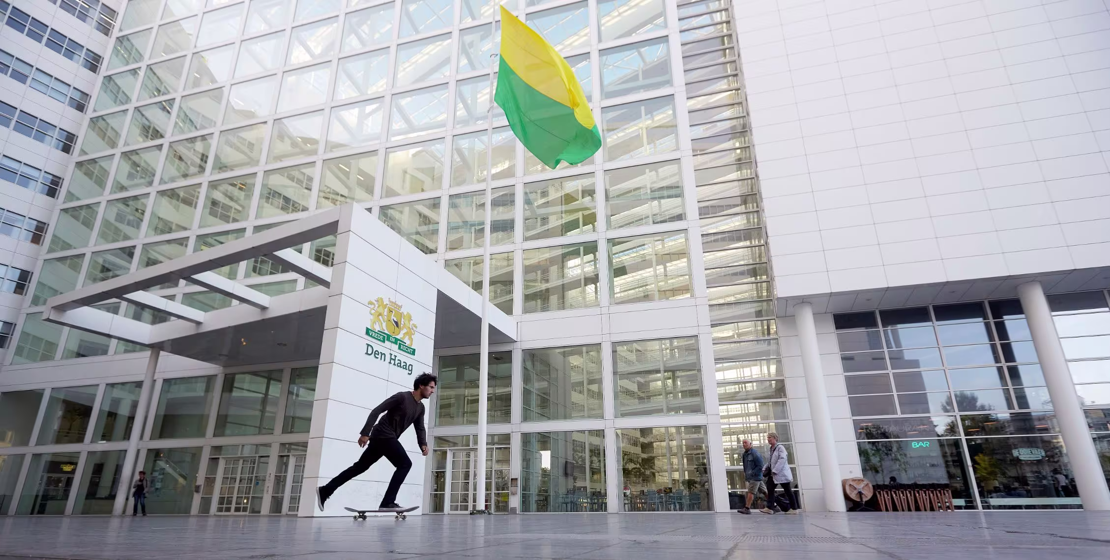 Skateboarder voor het moderne glazen gemeentehuis van Den Haag met geel-groene vlag.