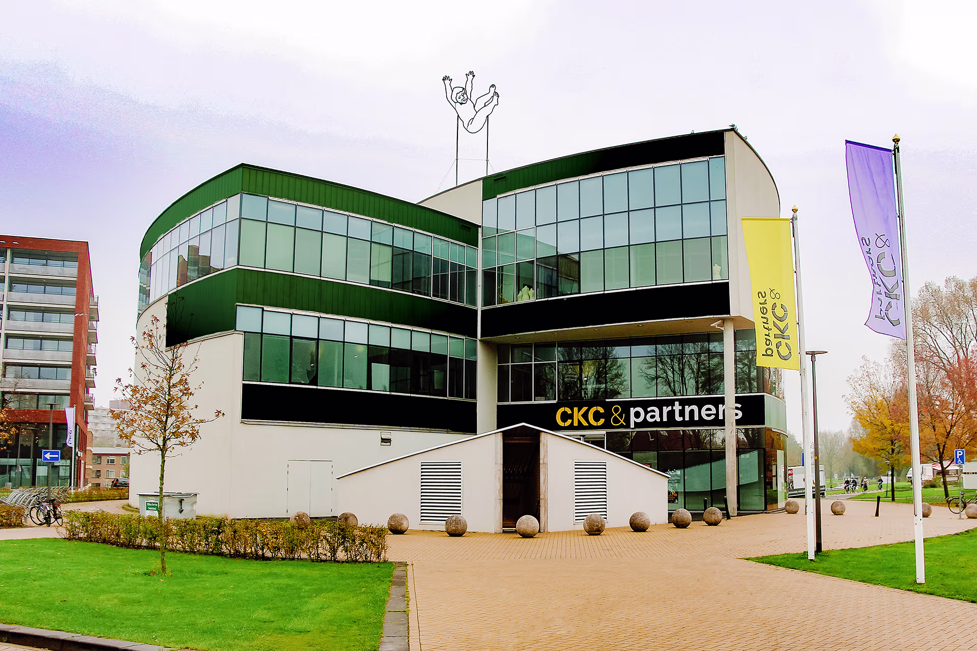 Modern kantoorgebouw van CKC & partners met groene en glazen gevel, omgeven door grasveld en gekleurde vlaggen.
