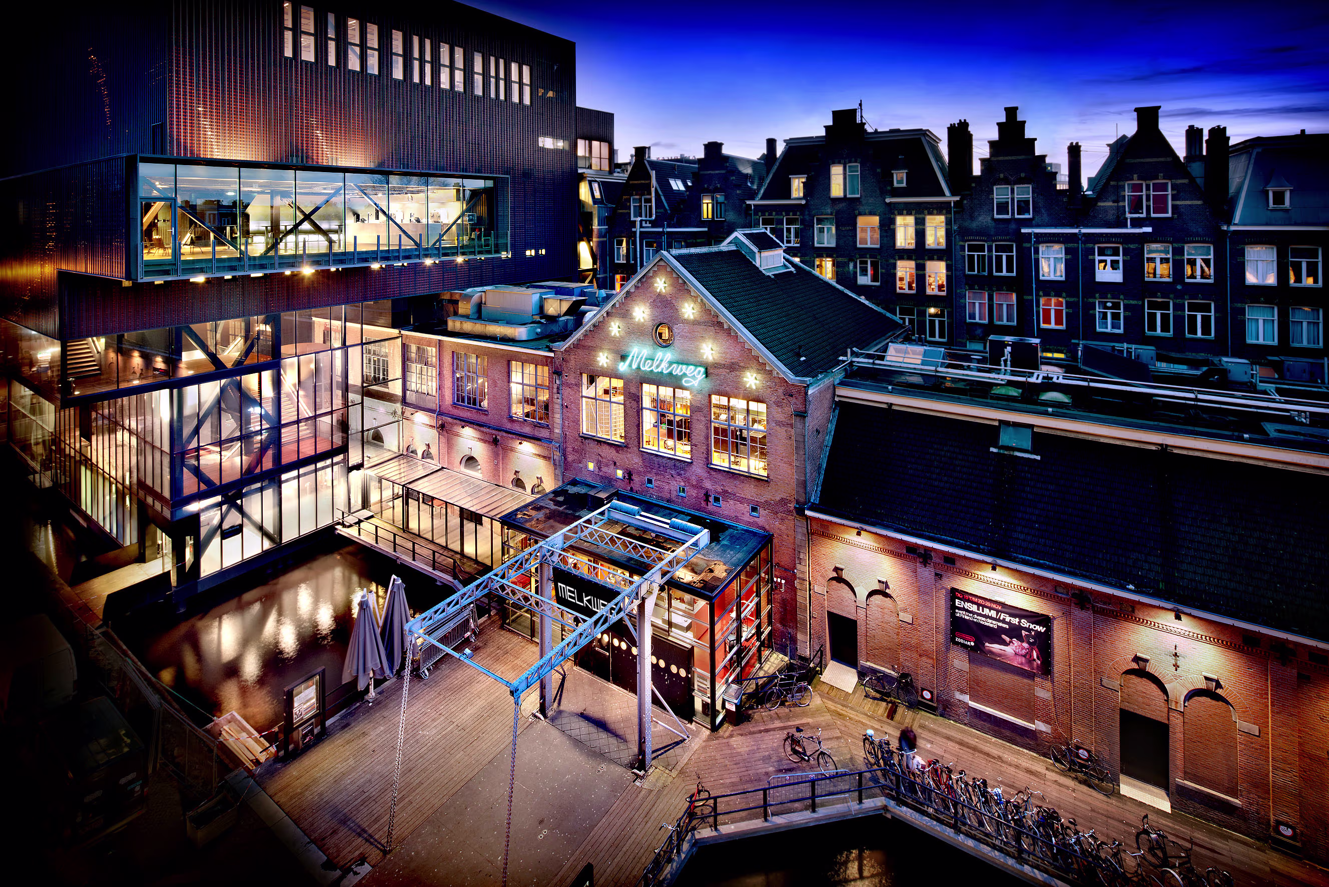 Nachtfoto van het moderne Melkweg cultuurgebouw in Amsterdam met verlichte ramen en traditionele grachtenhuizen op de achtergrond.