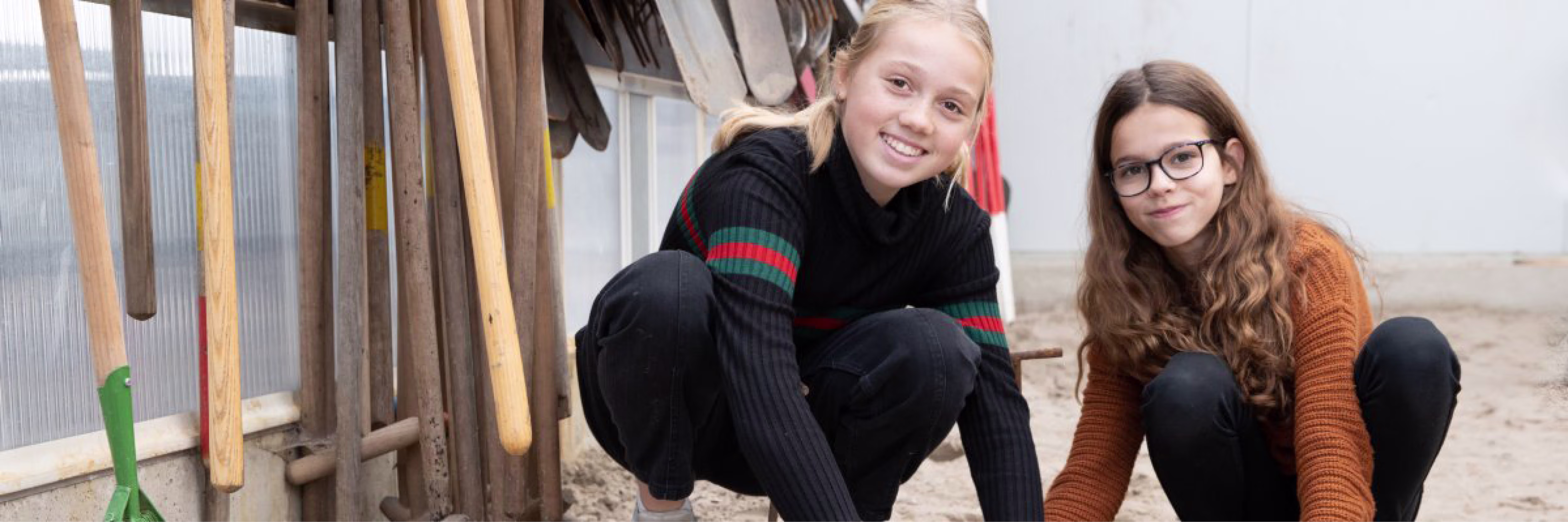 Twee jonge vrouwen zitten gehurkt bij houten palen op een strand en glimlachen naar de camera.