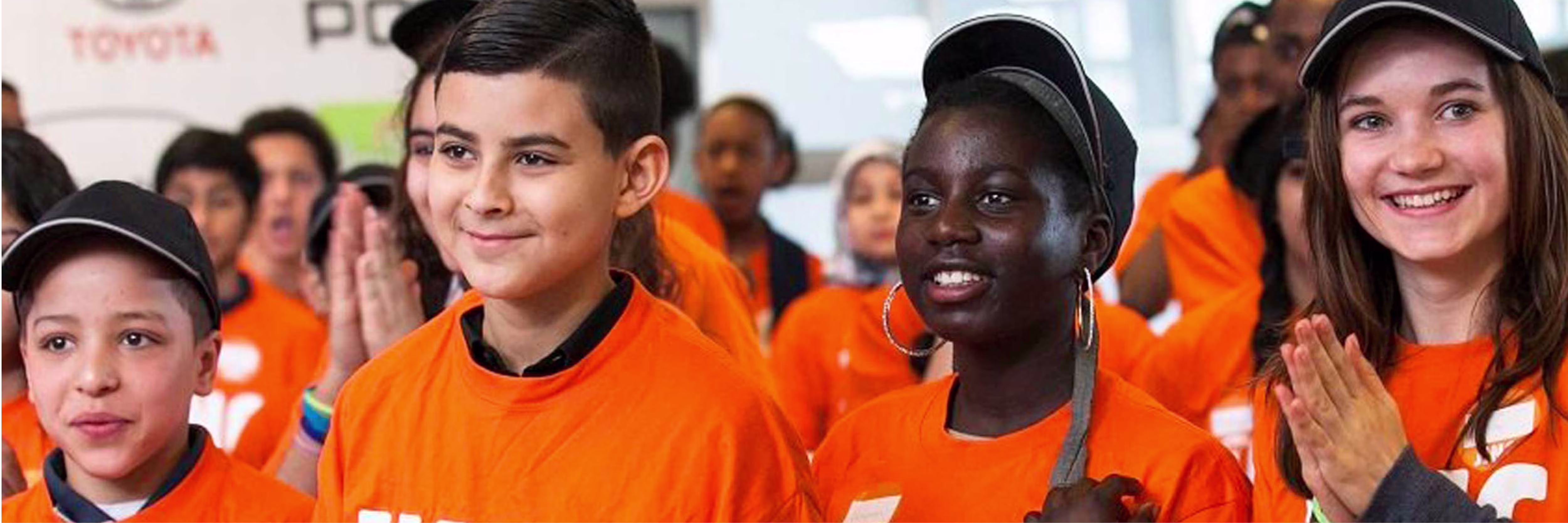 Vier jongeren in oranje shirts applaudisseren vrolijk tijdens een JINC evenement, met meer deelnemers op de achtergrond.