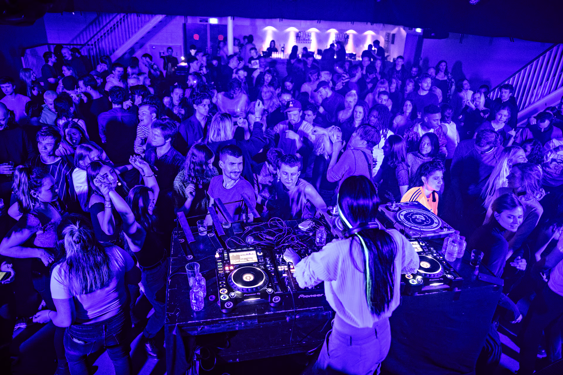 Een DJ draait muziek voor een dansende menigte in een club met paarse verlichting en twee verdiepingen.