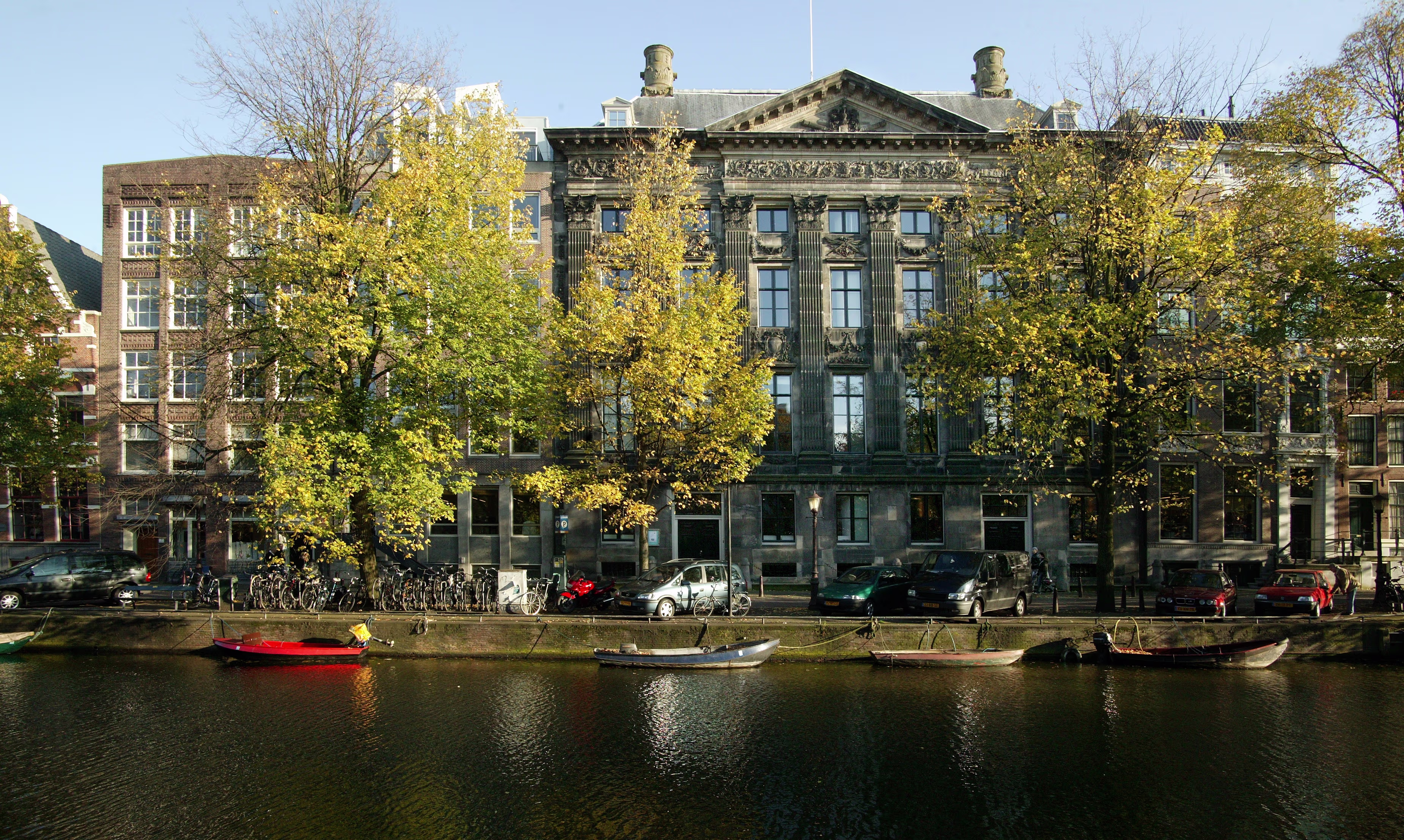 Historisch gebouw van de KNAW aan een Amsterdamse gracht met boten, omgeven door bomen en klassieke architectuur.