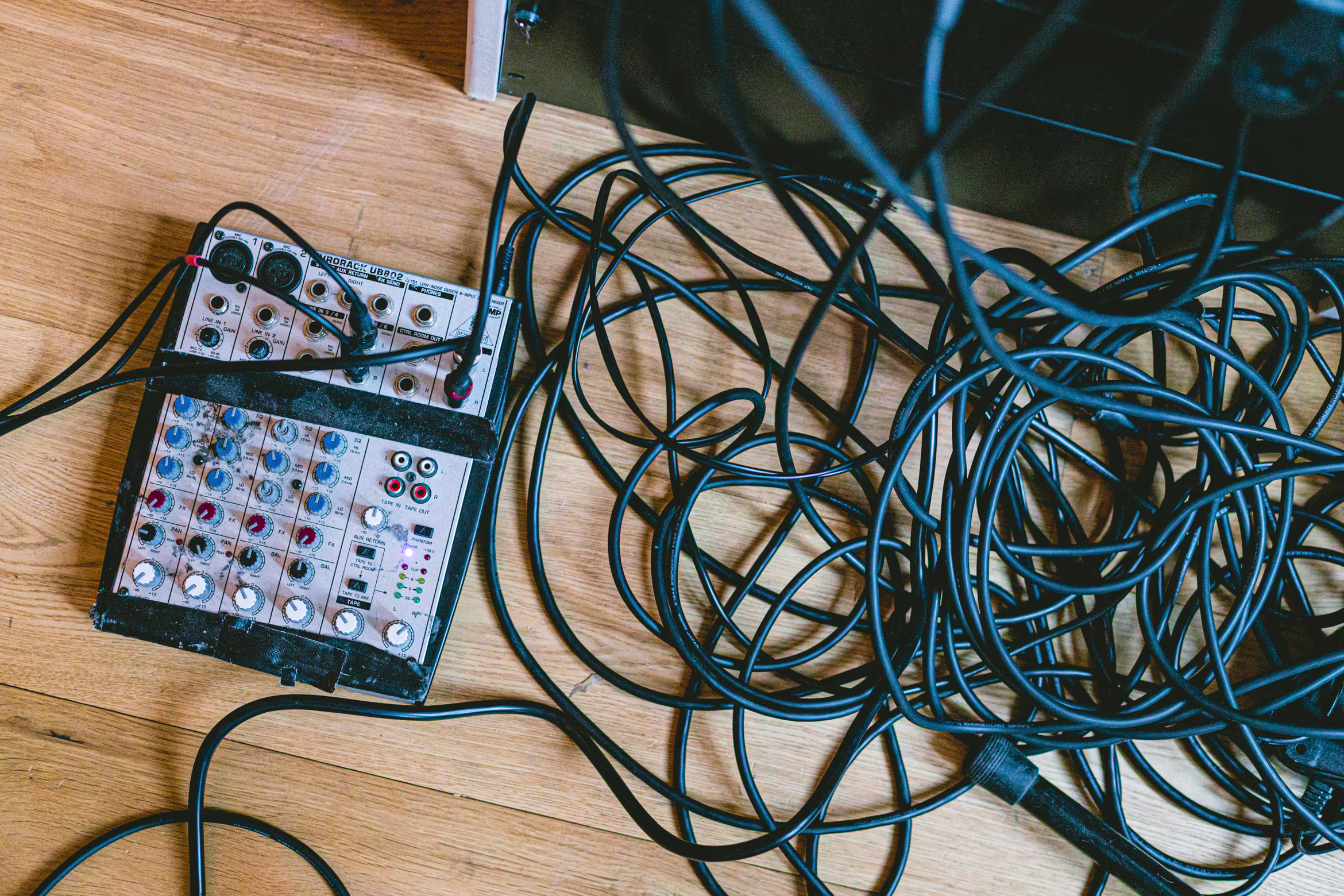 Een audiomixer met veel kabels ligt op een houten tafel, omgeven door een wirwar van zwarte snoeren.