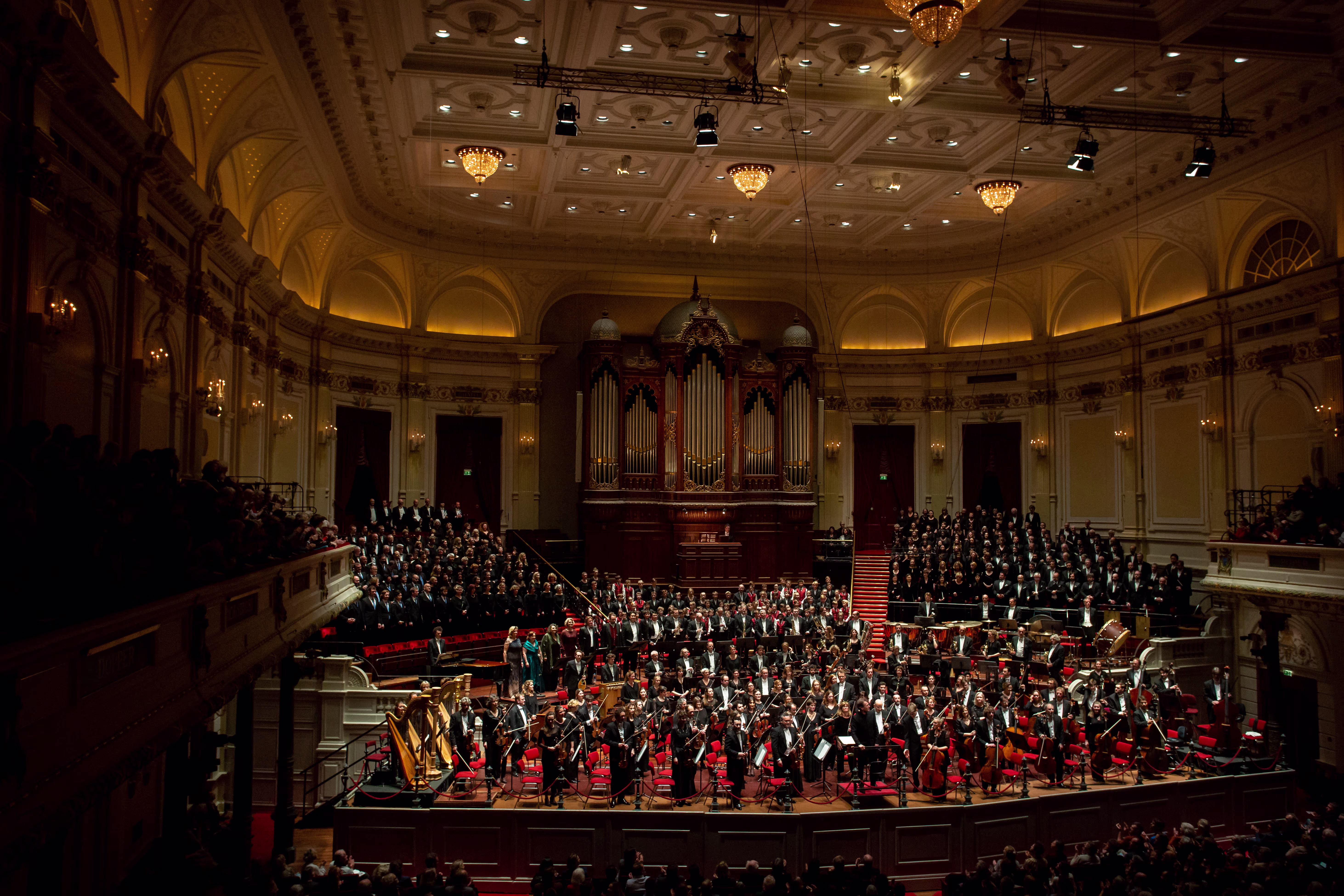 Een groot orkest en koor tijdens een uitvoering in een klassieke concerthal met orgel en publiek.