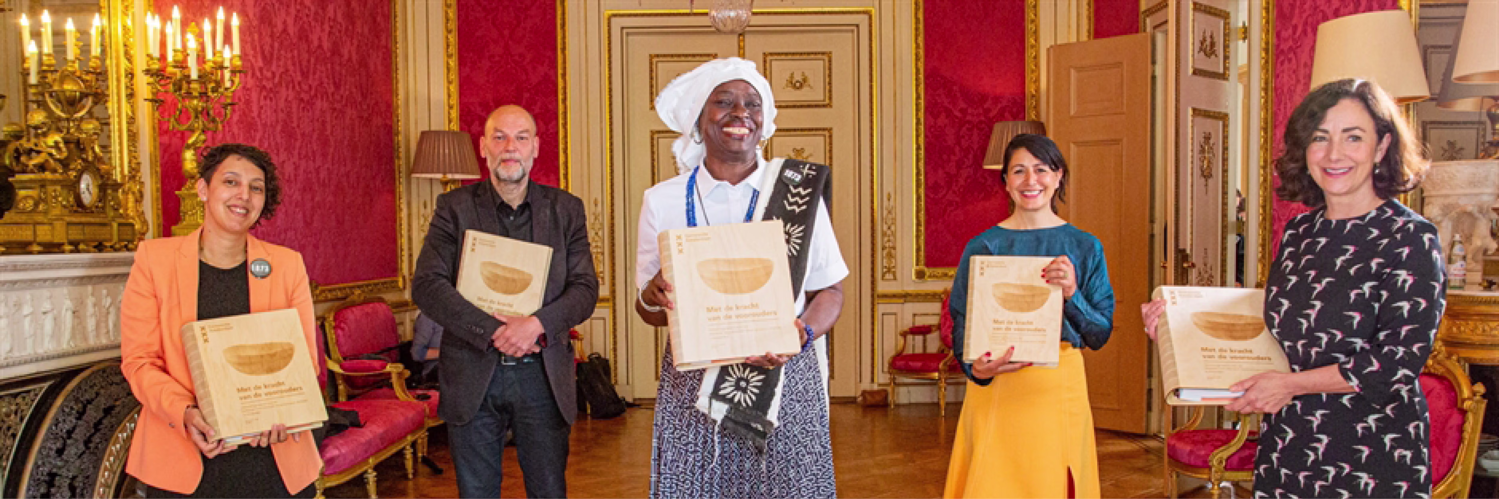 Vijf mensen in een elegante rode salon houden certificaten vast tijdens een officiële prijsuitreiking van de Gemeente Amsterdam.