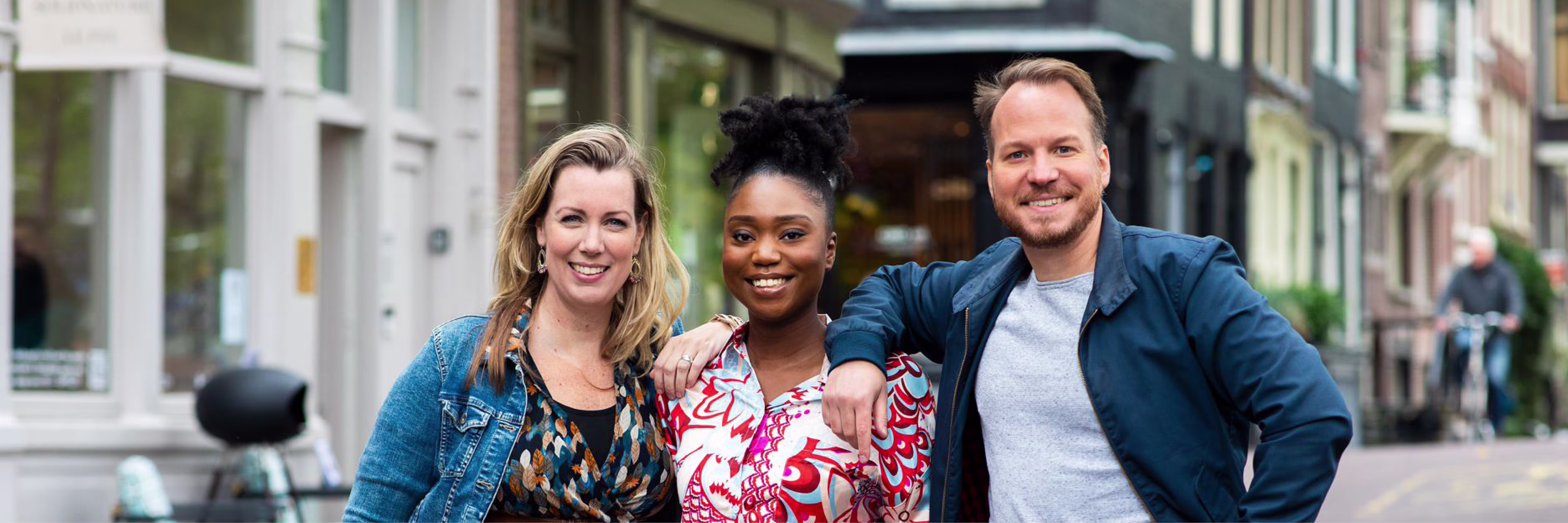 Drie glimlachende collega's staan arm in arm op een gezellige winkelstraat met historische panden op de achtergrond.