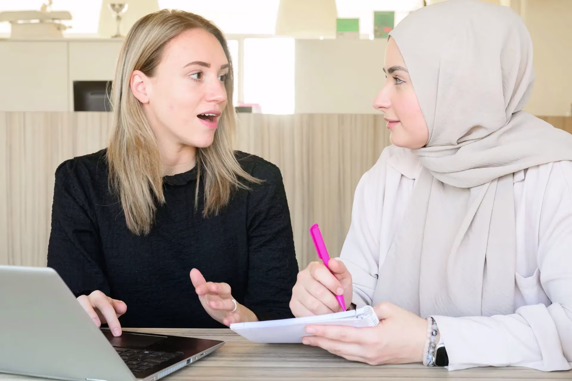 Twee vrouwen in gesprek aan bureau met laptop en notitieblok in kantooromgeving.