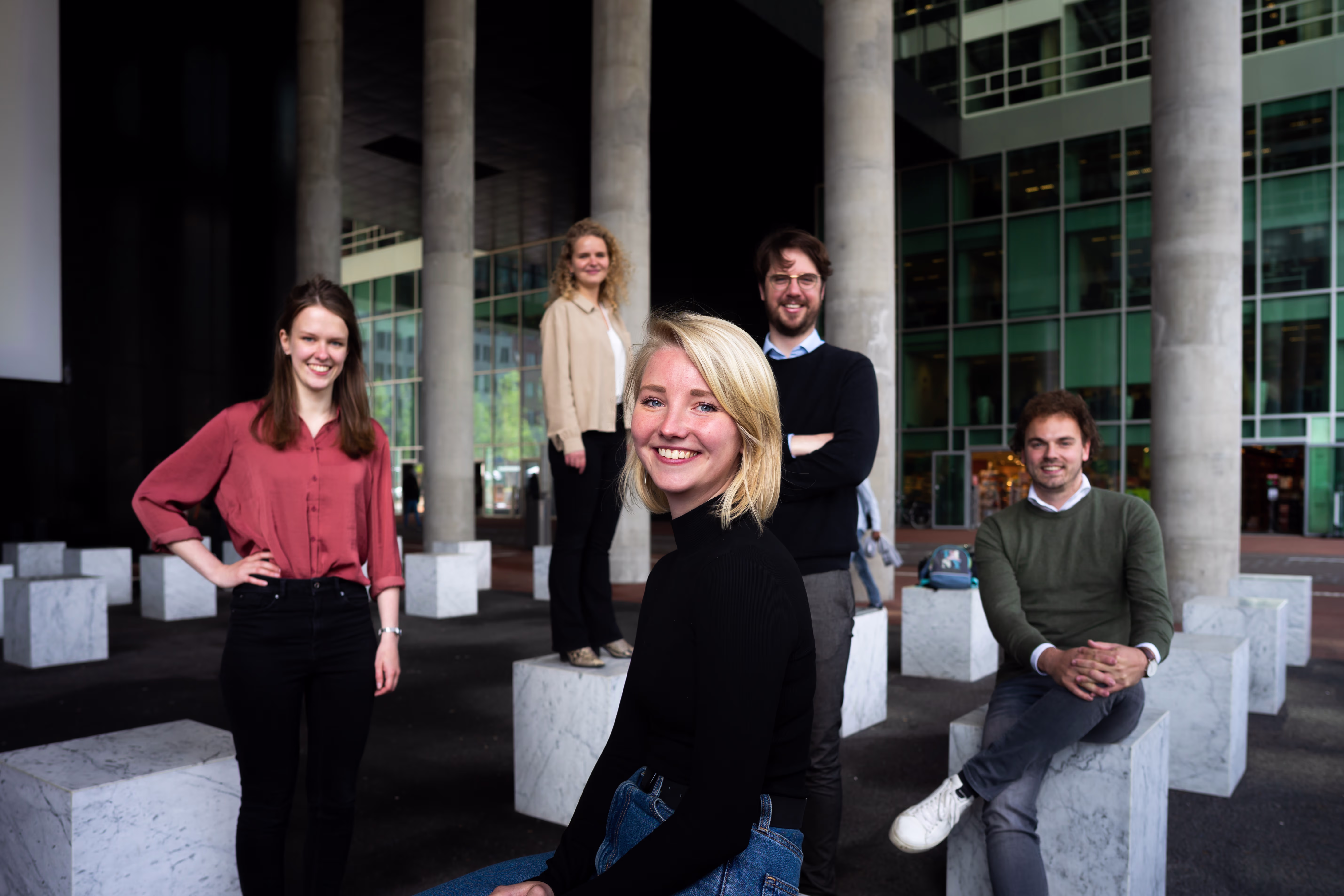 Five young professionals are posing in front of a modern office building with large glass windows and white columns.