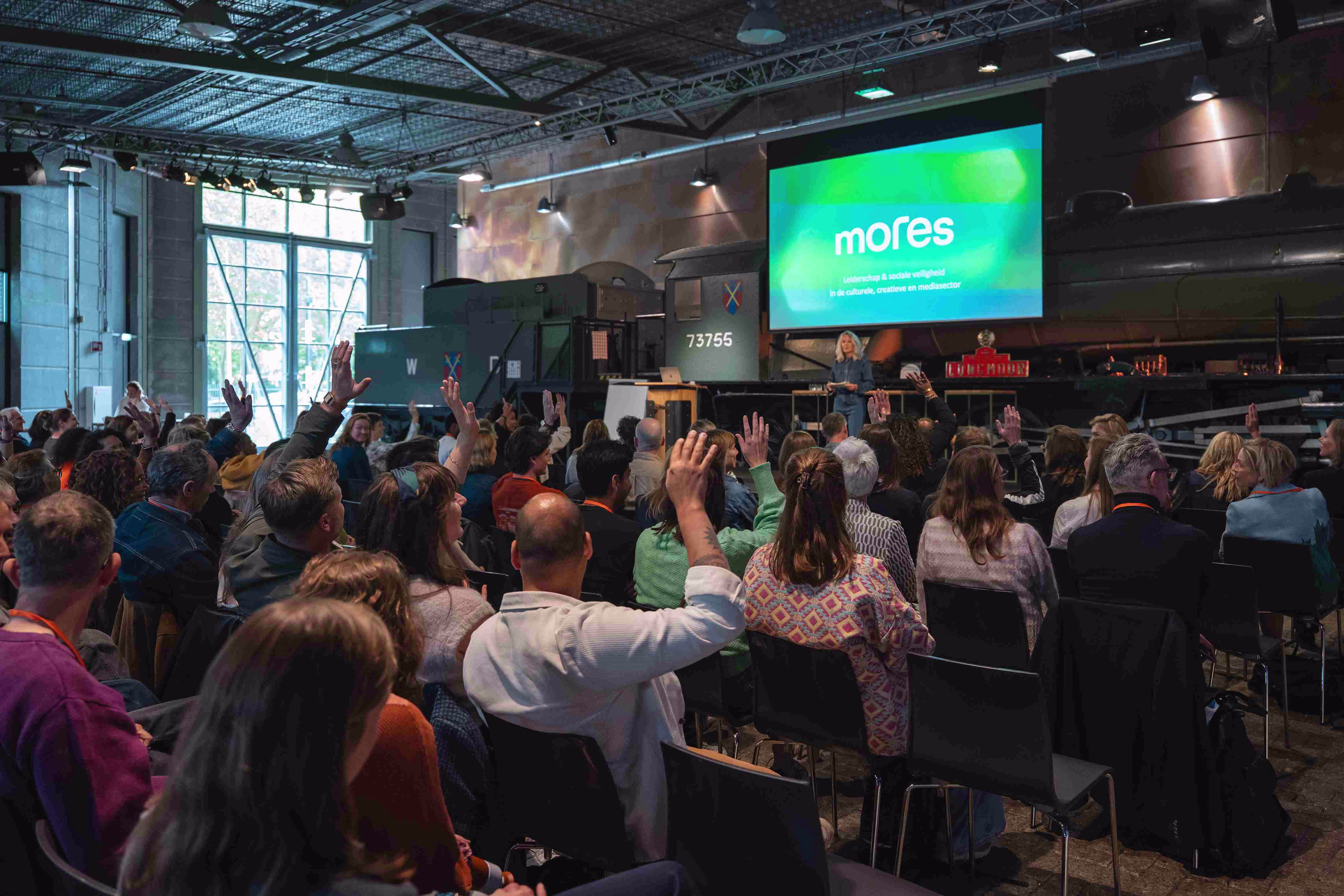 Een zaal vol mensen die hun hand opsteken tijdens een presentatie, met het Mores-logo op een groot scherm.