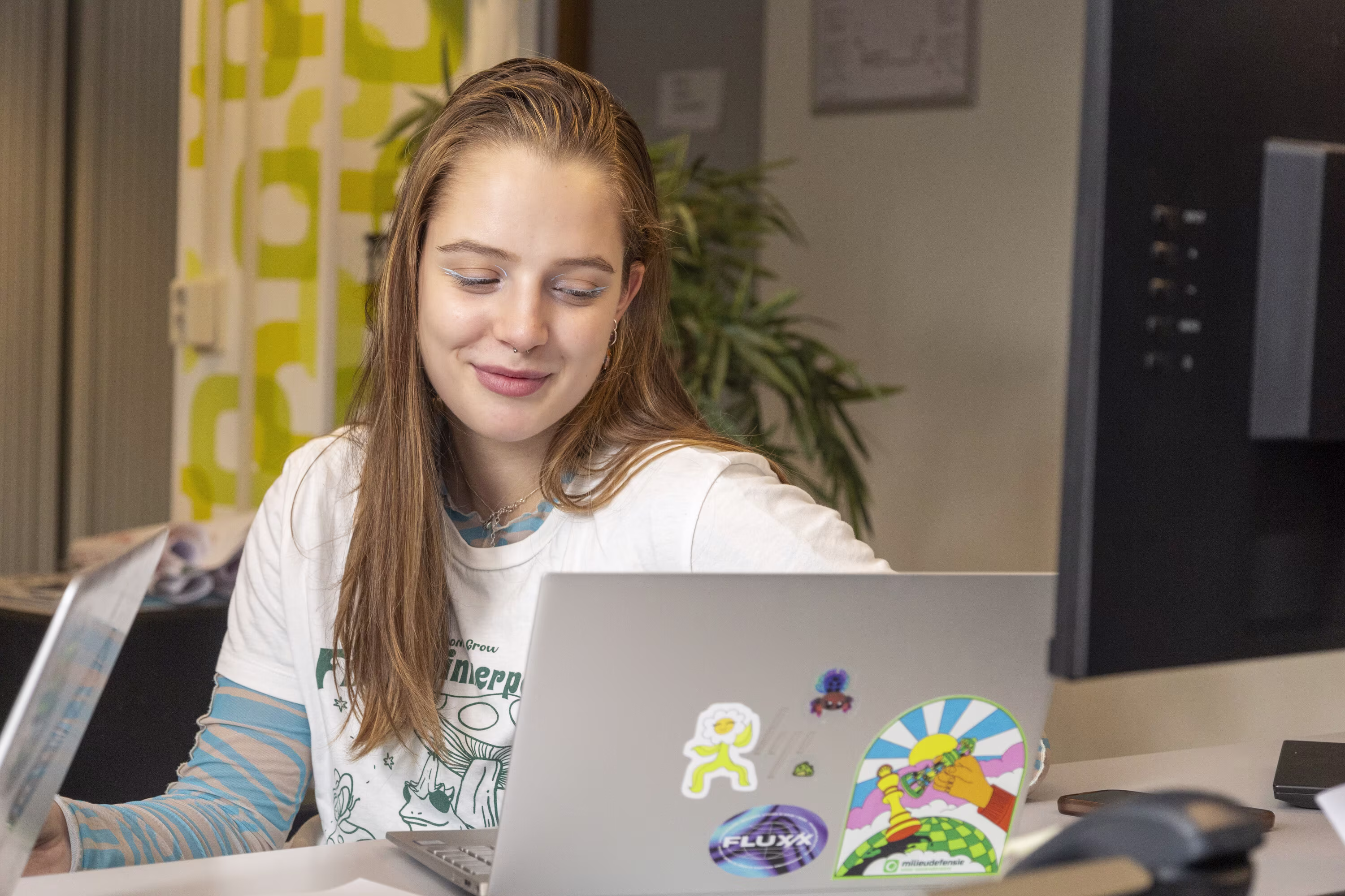Een jonge vrouw werkt glimlachend achter een laptop met kleurrijke stickers in een modern kantoor.