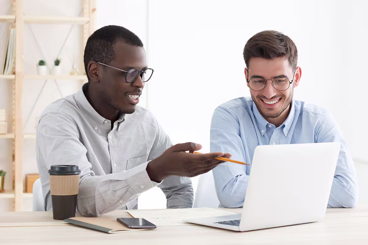 Twee lachende mannen in overhemd werken samen achter laptop op kantoor, een wijst naar het scherm.