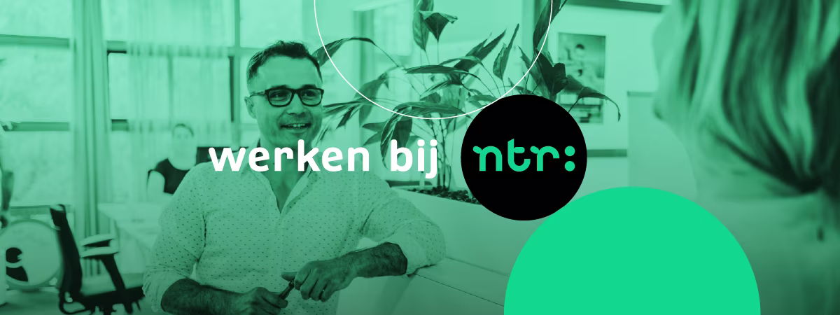 Een man met bril zit lachend aan een bureau in een kantooromgeving met planten op de achtergrond en het NTR-logo.