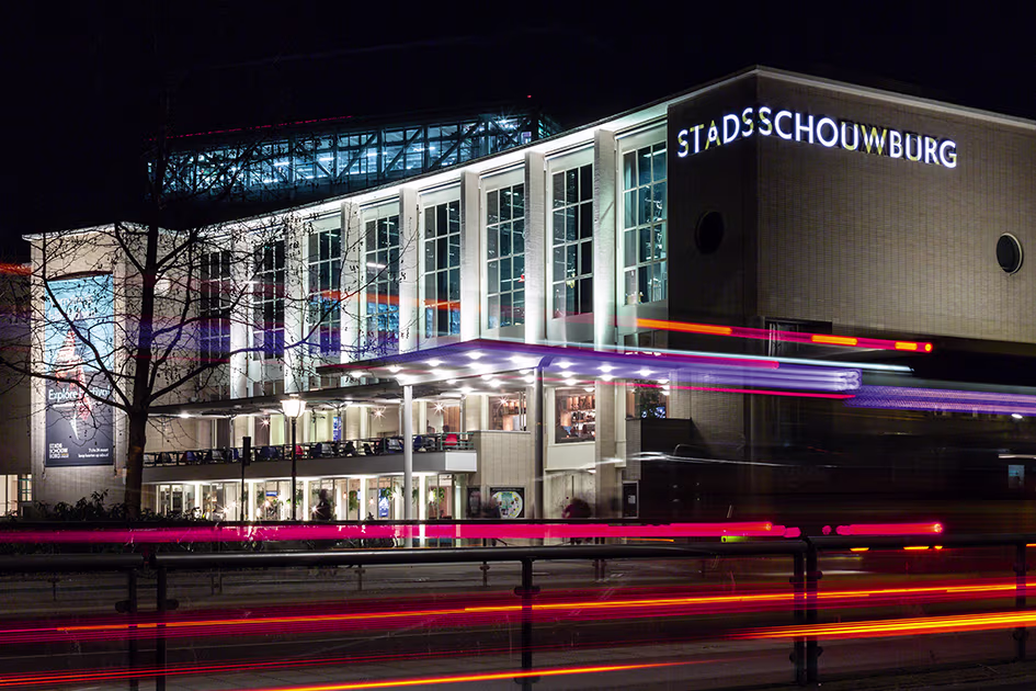 Stadsschouwburg Utrecht verlicht in de avond met kleurrijke lichtstrepen van langsrijdend verkeer op de voorgrond.