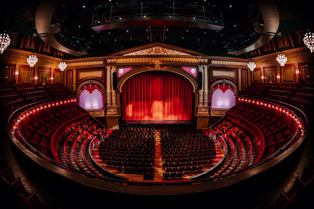 Luxueuze theaterzaal met rode gordijnen, kristallen kroonluchters en rijen stoelen in een cirkelvormige opstelling.