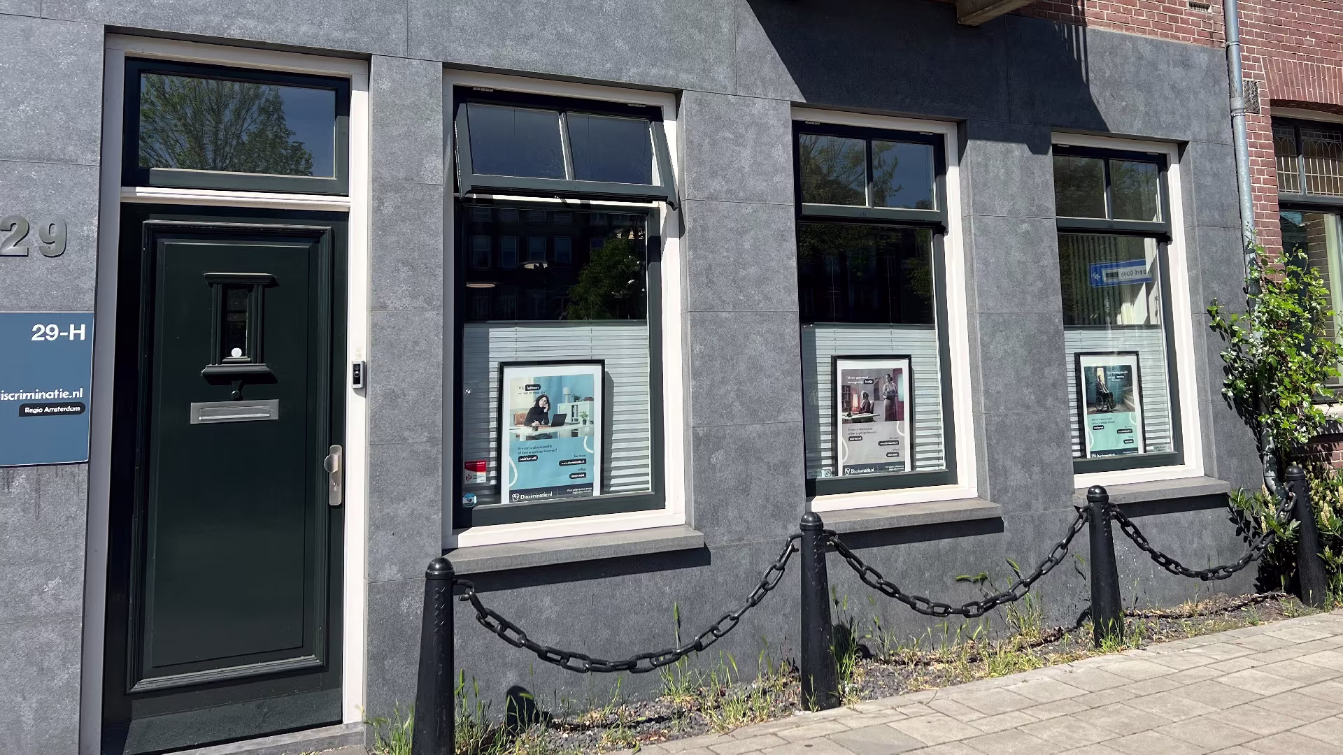 A gray commercial building with a green front door, number 29-H, and three display windows featuring posters from Discriminatie.nl Amsterdam Region.