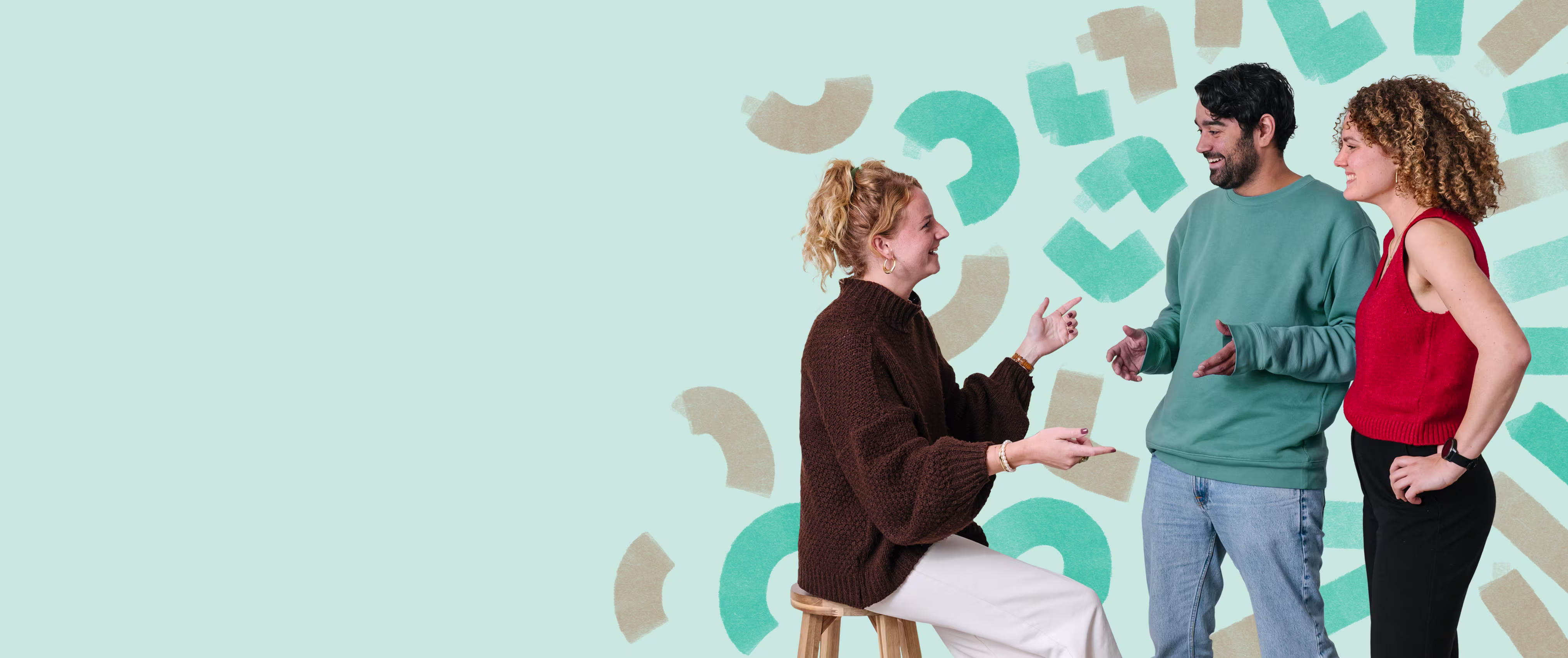 Three colleagues are talking in front of a wall with green and beige shapes; one is sitting on a stool, and two are standing.