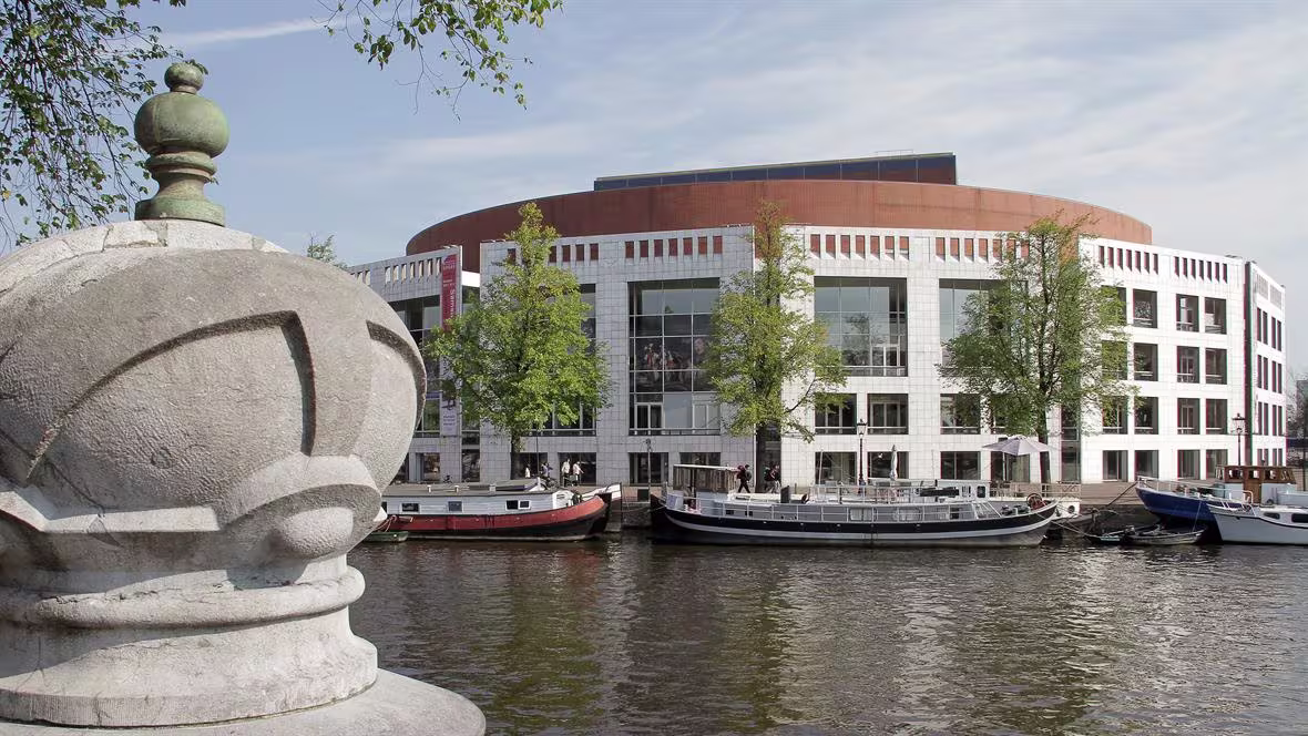 Modern gemeentehuis aan Amsterdamse gracht met historische boot en decoratief hekwerk op de voorgrond.