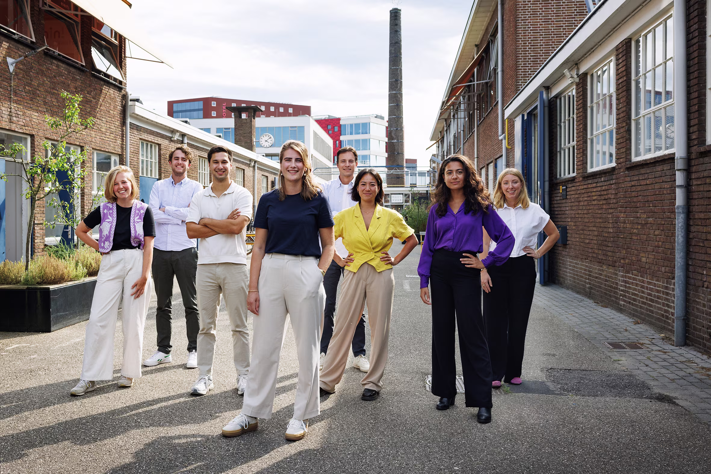 A group of eight people dressed in business attire poses together on the street between modern office buildings.