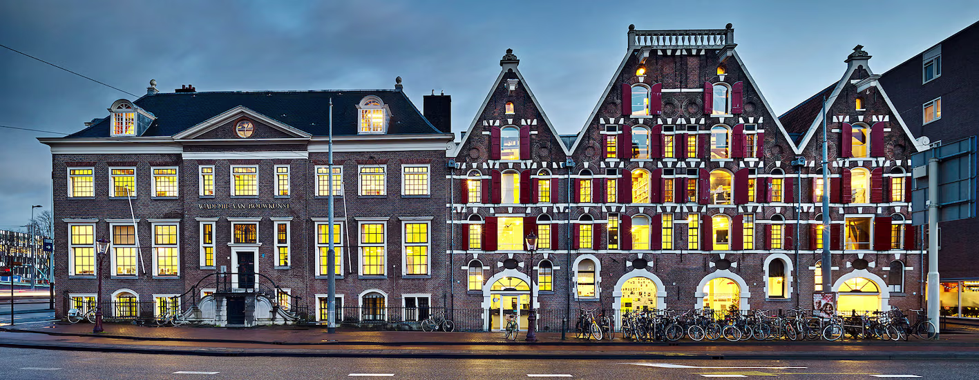 Historische gebouwen van de Academie van Bouwkunst Amsterdam met verlichte ramen tijdens de avondschemering.