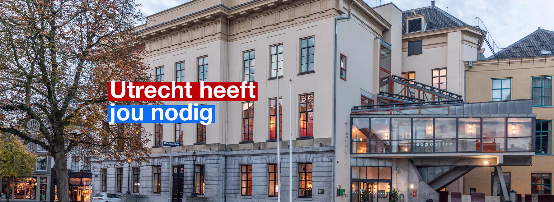 Historisch gebouw van de Gemeente Utrecht met moderne glazen uitbouw, verlicht in de avondschemering.