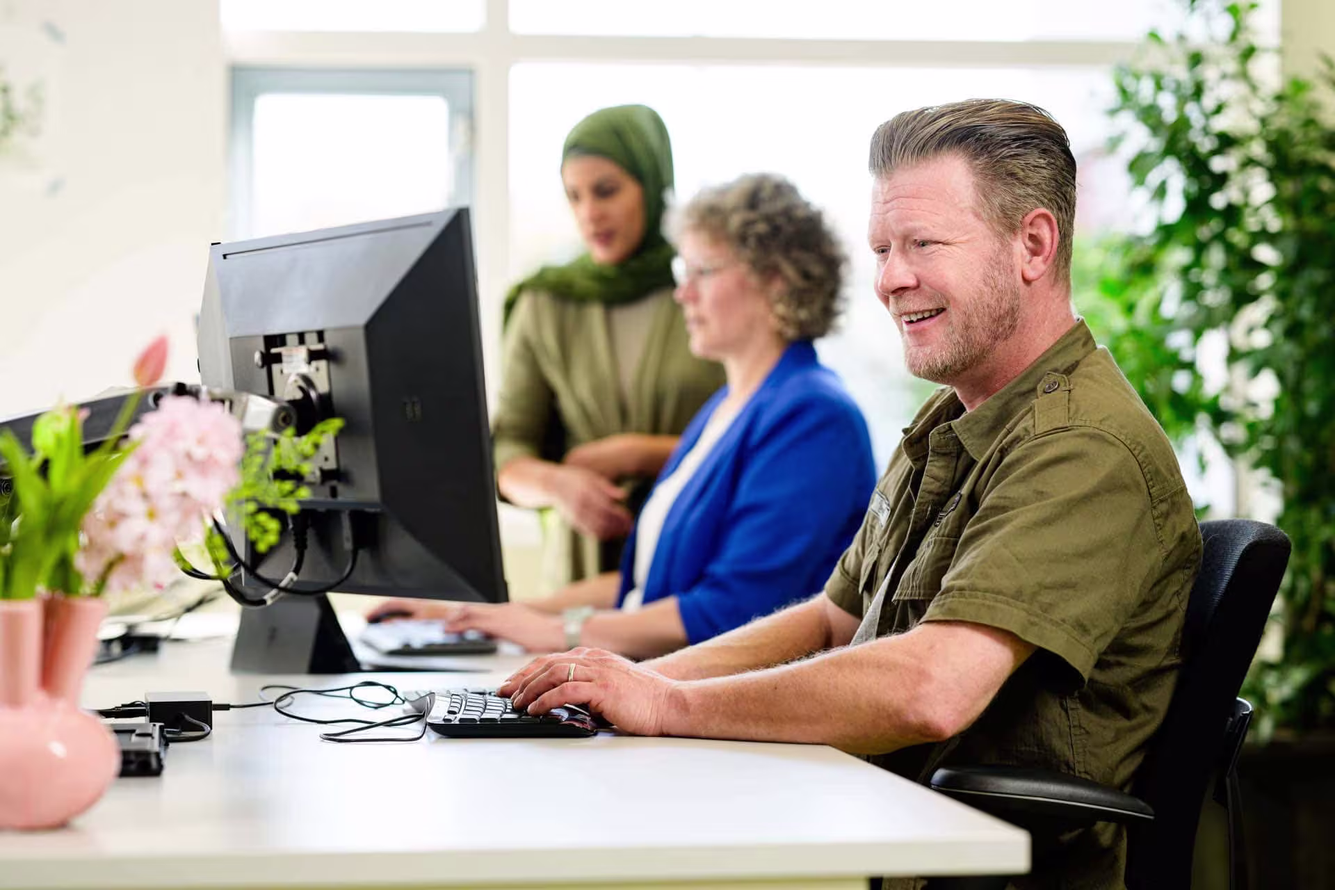 Lachende man in groen shirt werkt aan computer in modern kantoor, collega's op achtergrond, planten en bloemen zichtbaar.