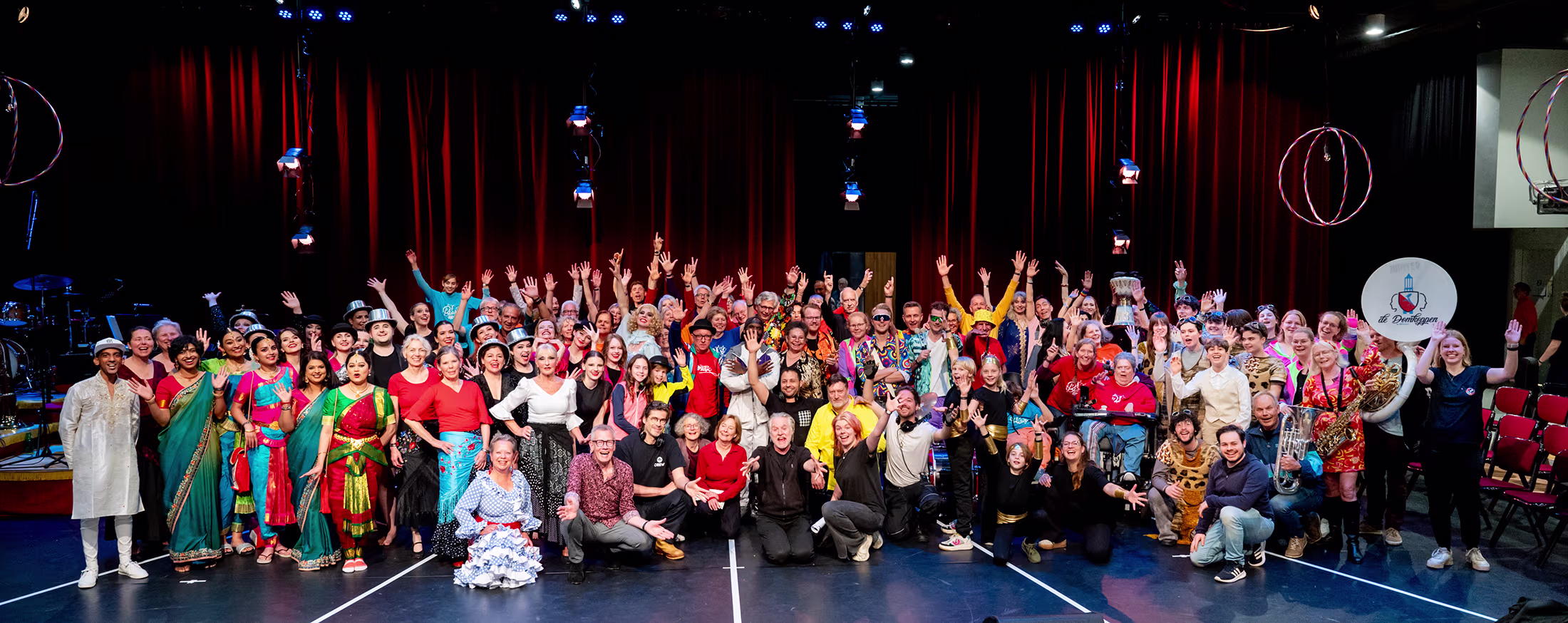 Grote groep mensen in kleurrijke kostuums poseert samen op een podium met rode gordijnen na een theatrale voorstelling.