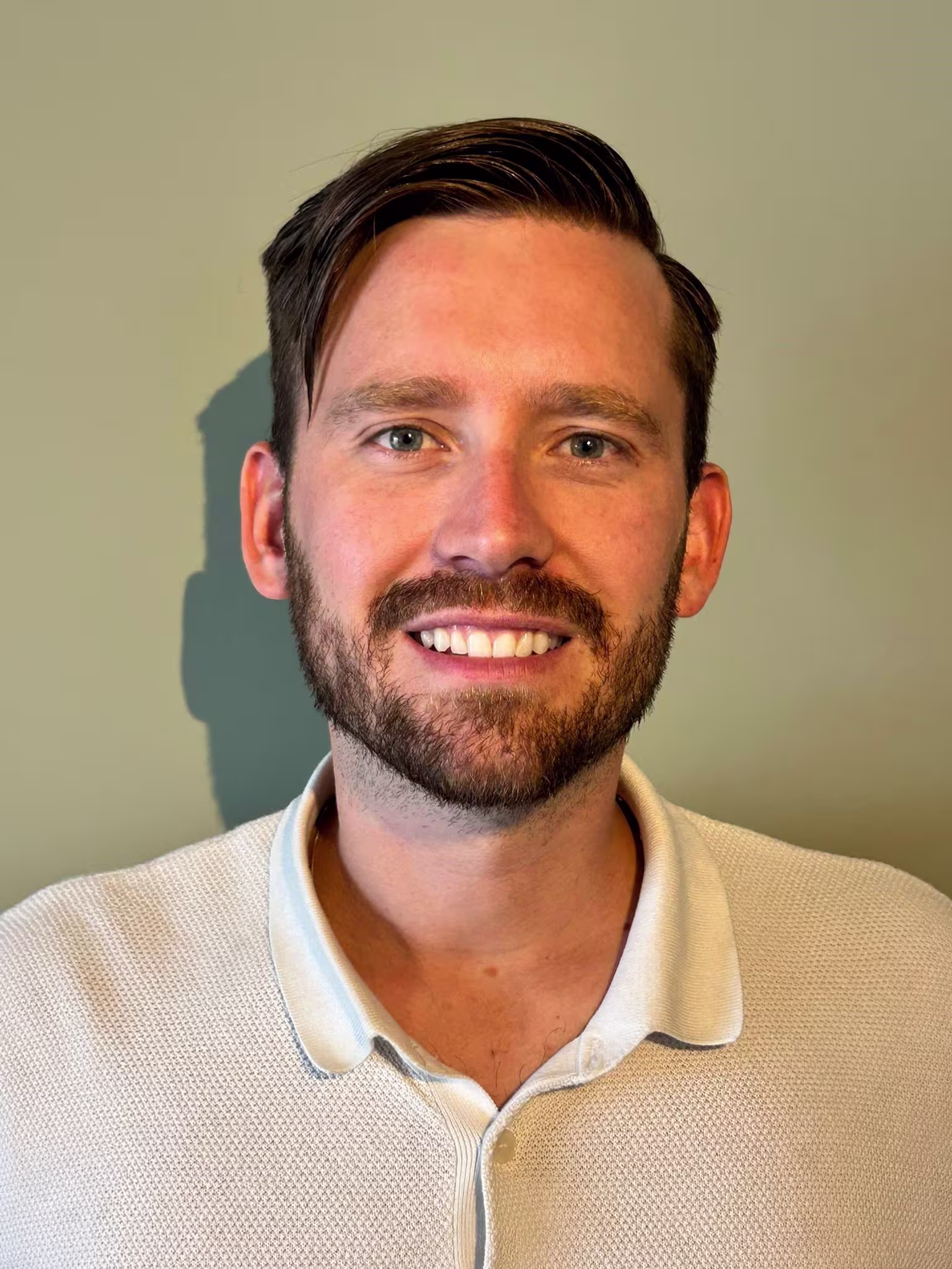 Glimlachende man met baard in wit overhemd poseert voor professionele portretfoto tegen neutrale achtergrond.