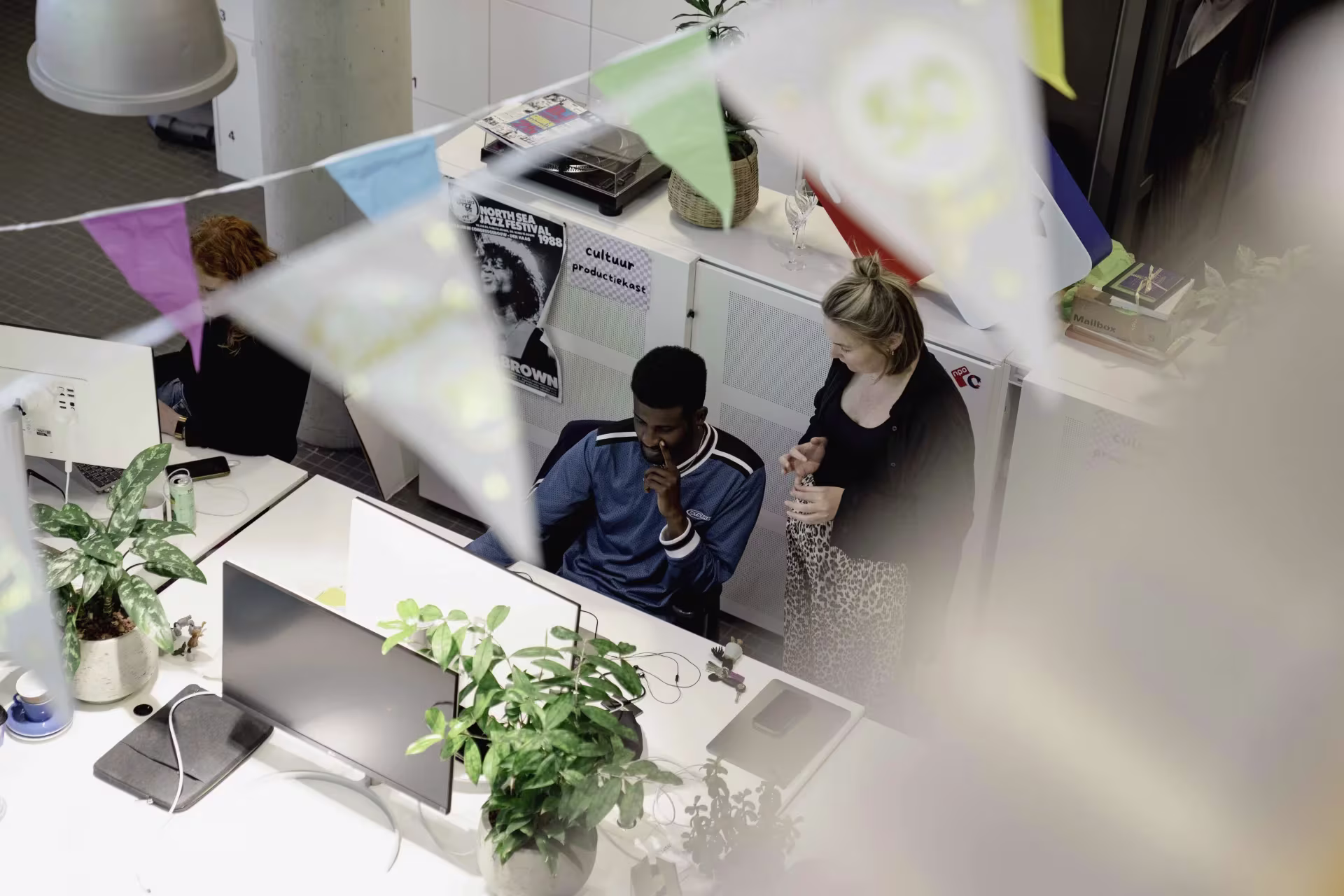 Two employees talking at a desk in a modern office decorated with plants and colorful accents.