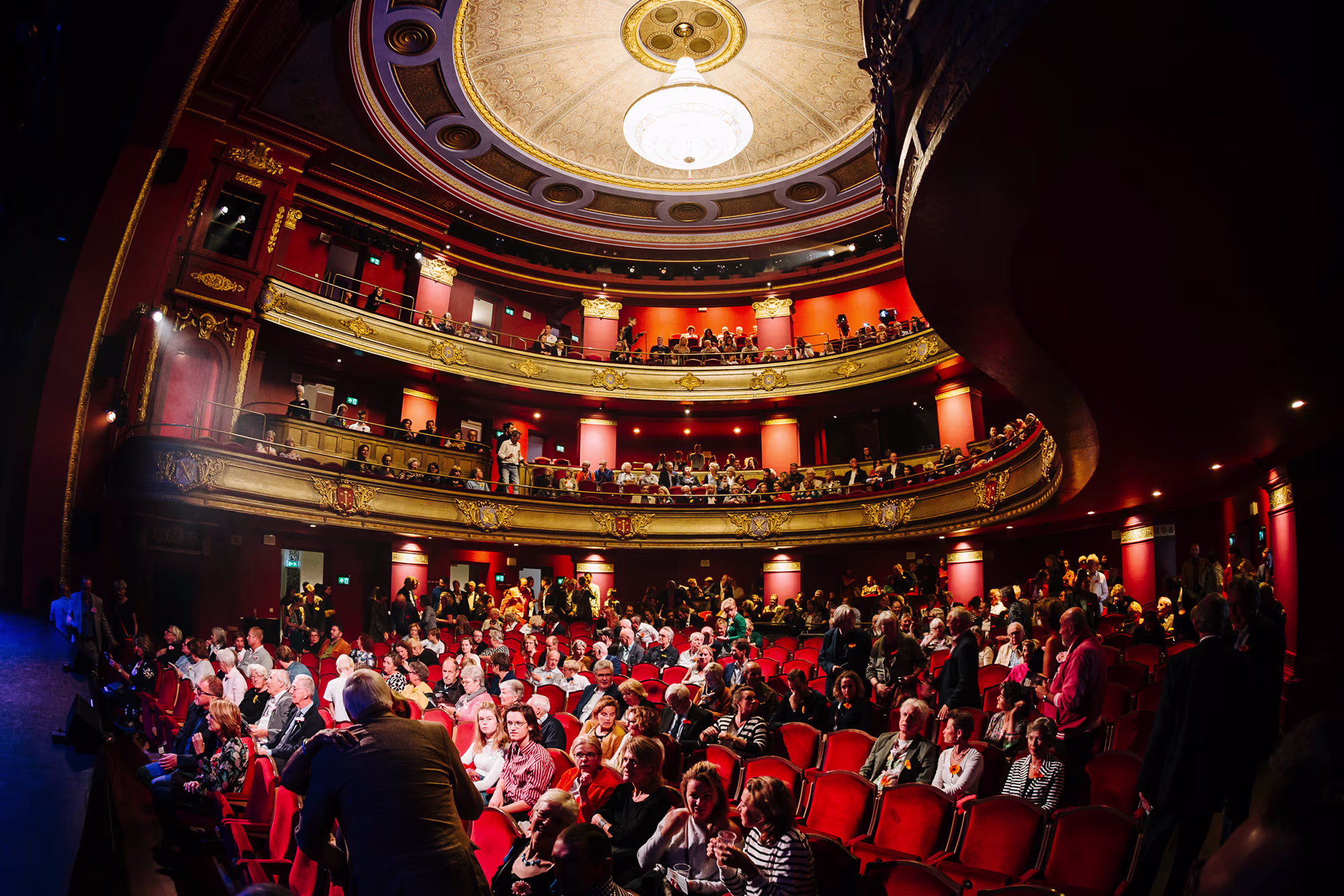 Publiek in een traditionele schouwburg met rode stoelen, gouden balkons en een koepelplafond tijdens een voorstelling.