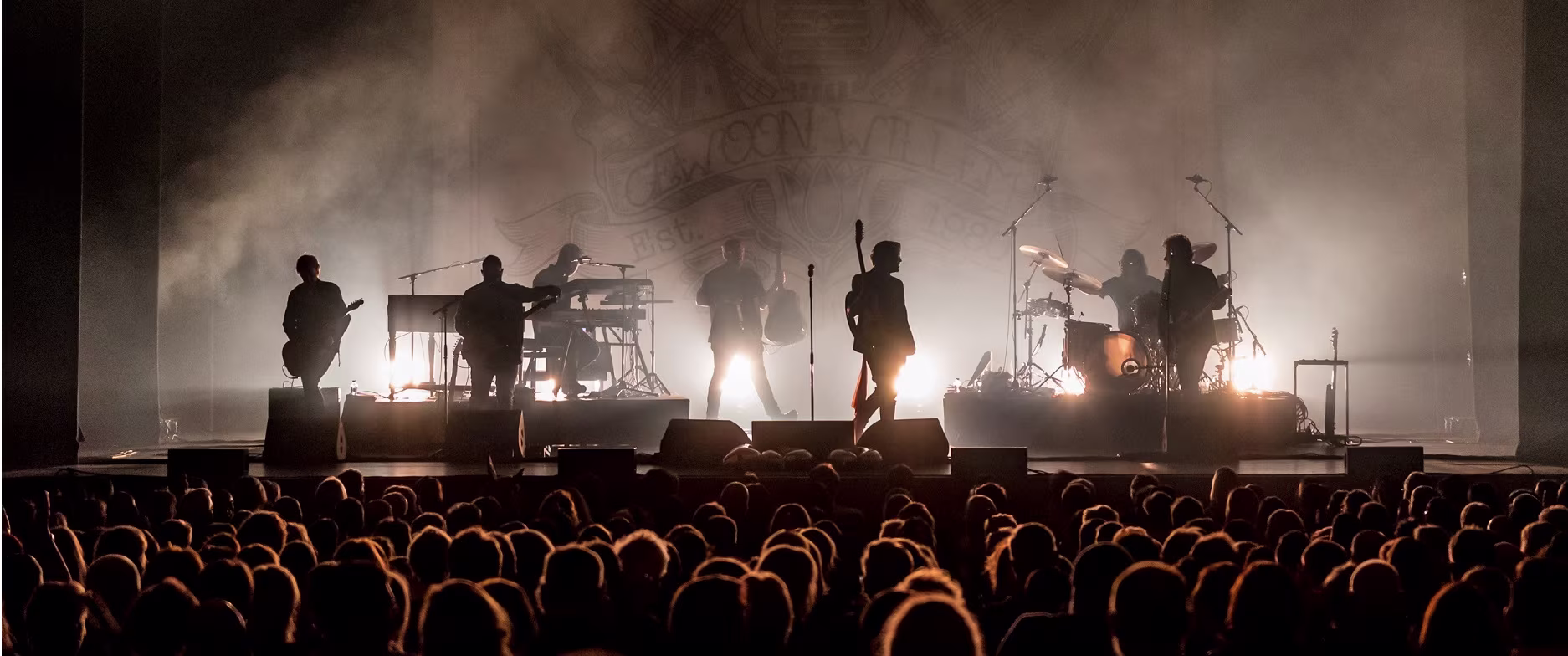 Een band treedt op voor een groot publiek in een theater, met dramatische belichting en rookeffecten op het podium.