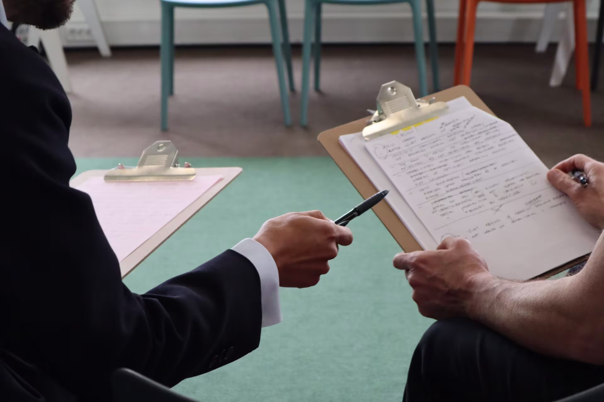 Twee personen in pak bespreken documenten op klemborden aan een tafel in een moderne kantooromgeving.