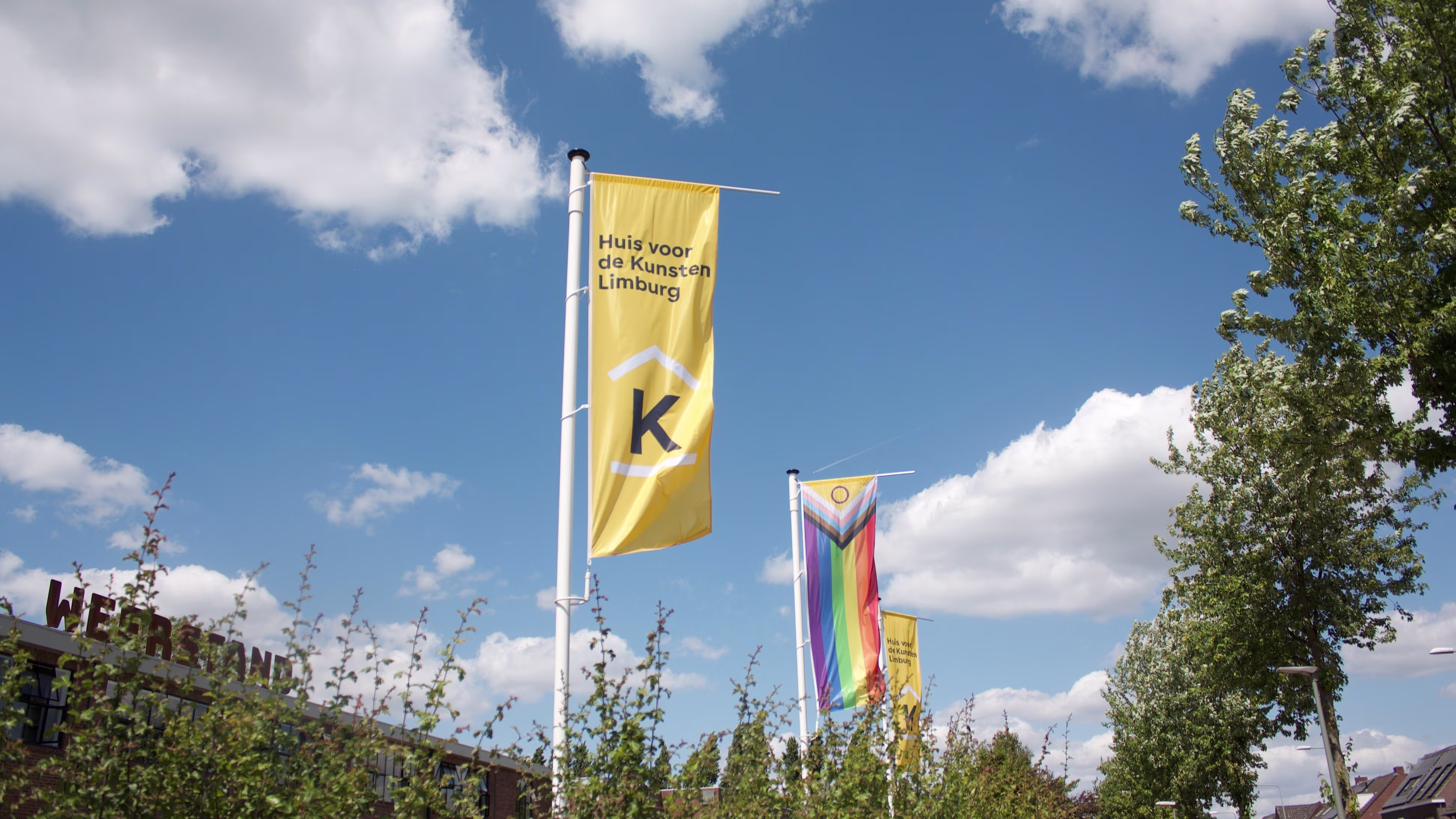 Gele vlaggen met logo Huis voor de Kunsten Limburg en regenboogvlag wapperen tegen blauwe hemel met wolken.
