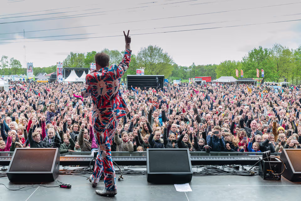 Een performer in kleurrijk pak staat op een podium voor een grote, juichende menigte tijdens een festival.