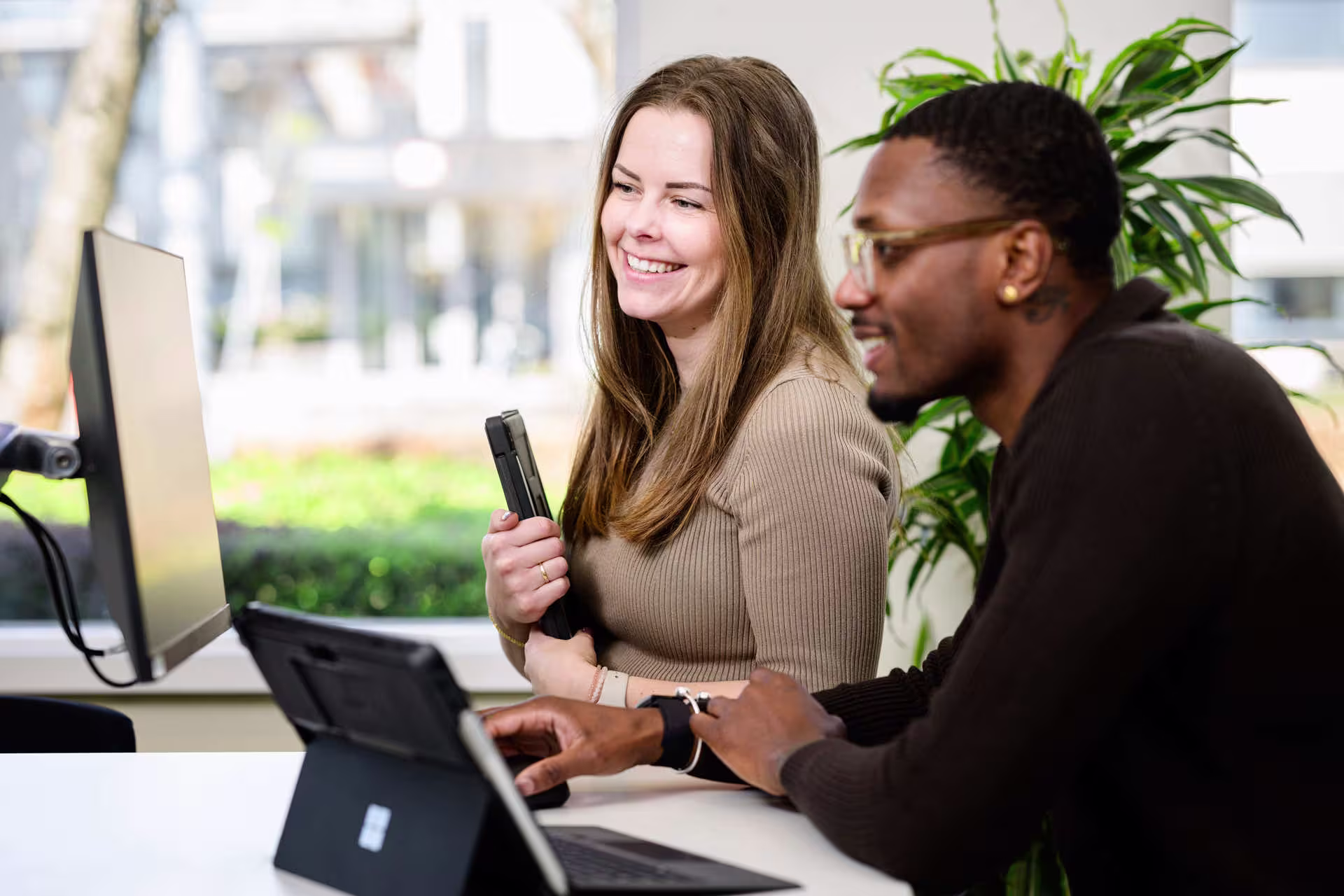 Twee collega's werken samen achter laptops in een modern kantoor met planten op de achtergrond.