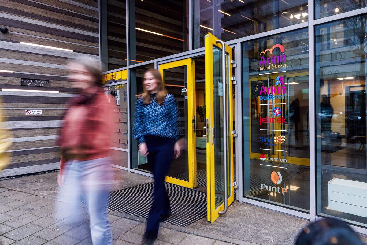 Twee personen lopen door de gele toegangsdeur van een modern glazen gebouw met het logo van Arkin zichtbaar op de gevel.