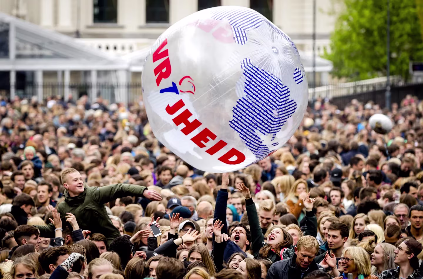 Grote menigte mensen op een plein, iemand gooit een witte bal met tekst omhoog, vrolijke sfeer bij een evenement.