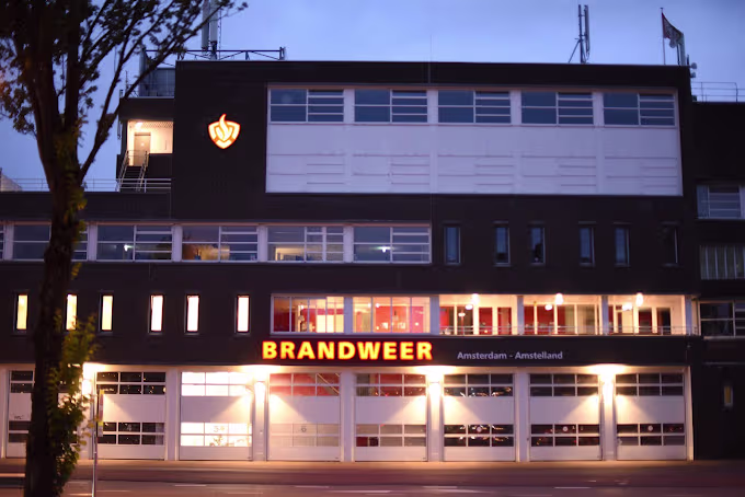 Brandweerkazerne van Veiligheidsregio Amsterdam-Amstelland bij avond, modern gebouw met verlichte ramen en garagedeuren.