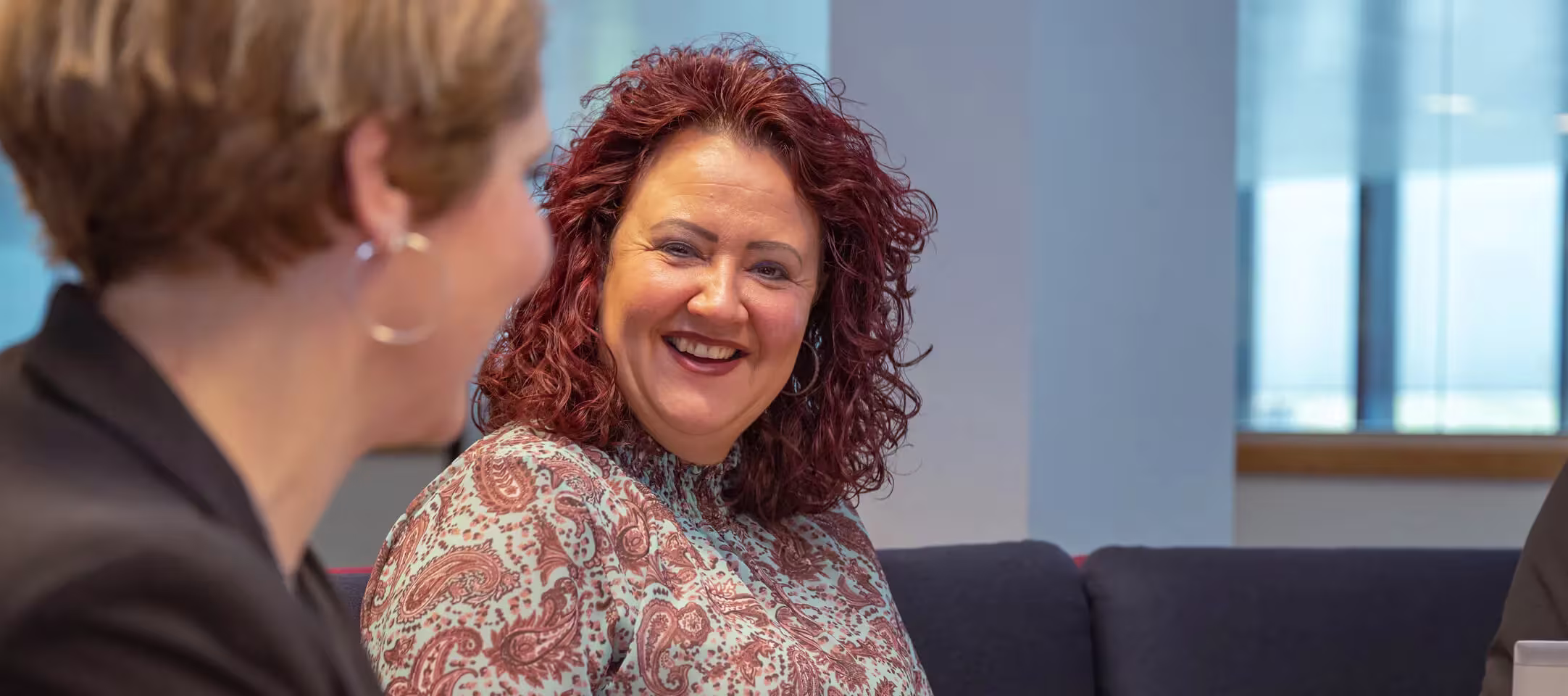 Two women chatting in the office; one is smiling cheerfully at the other in a modern workspace with large windows.