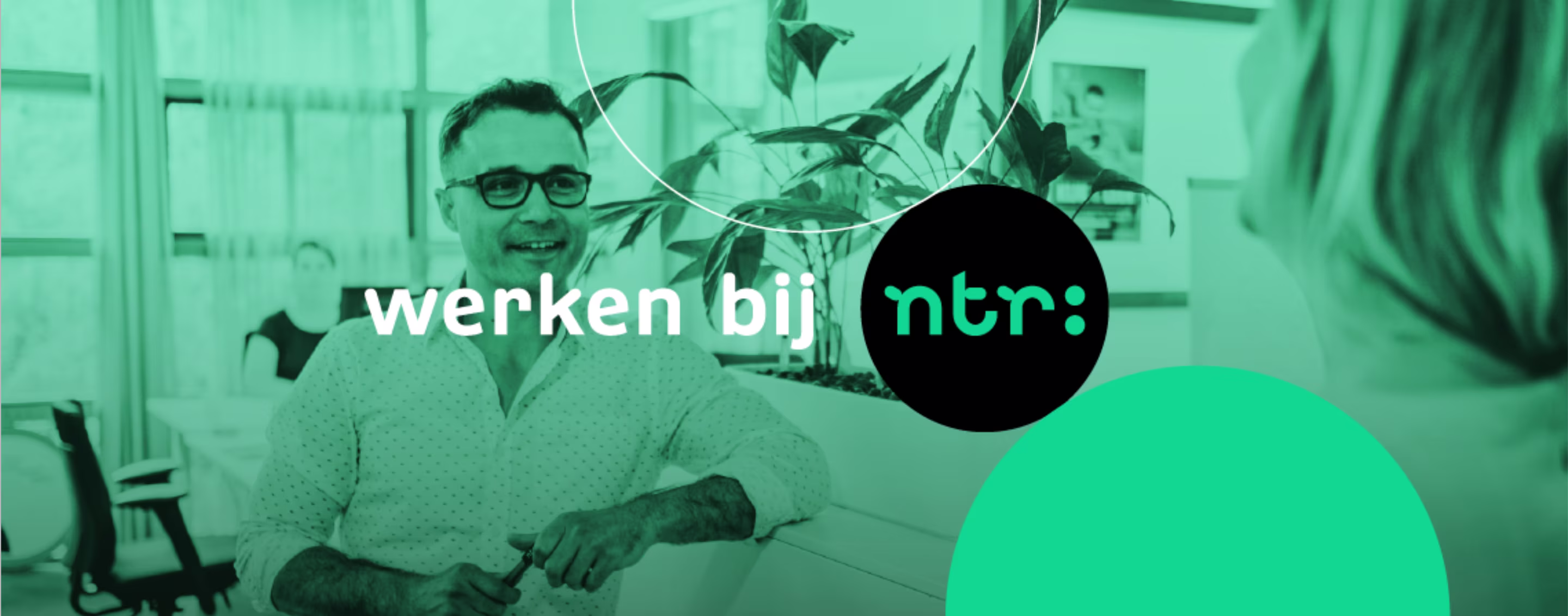Een lachende man met bril zit aan een bureau in een moderne kantooromgeving met planten, met het logo 